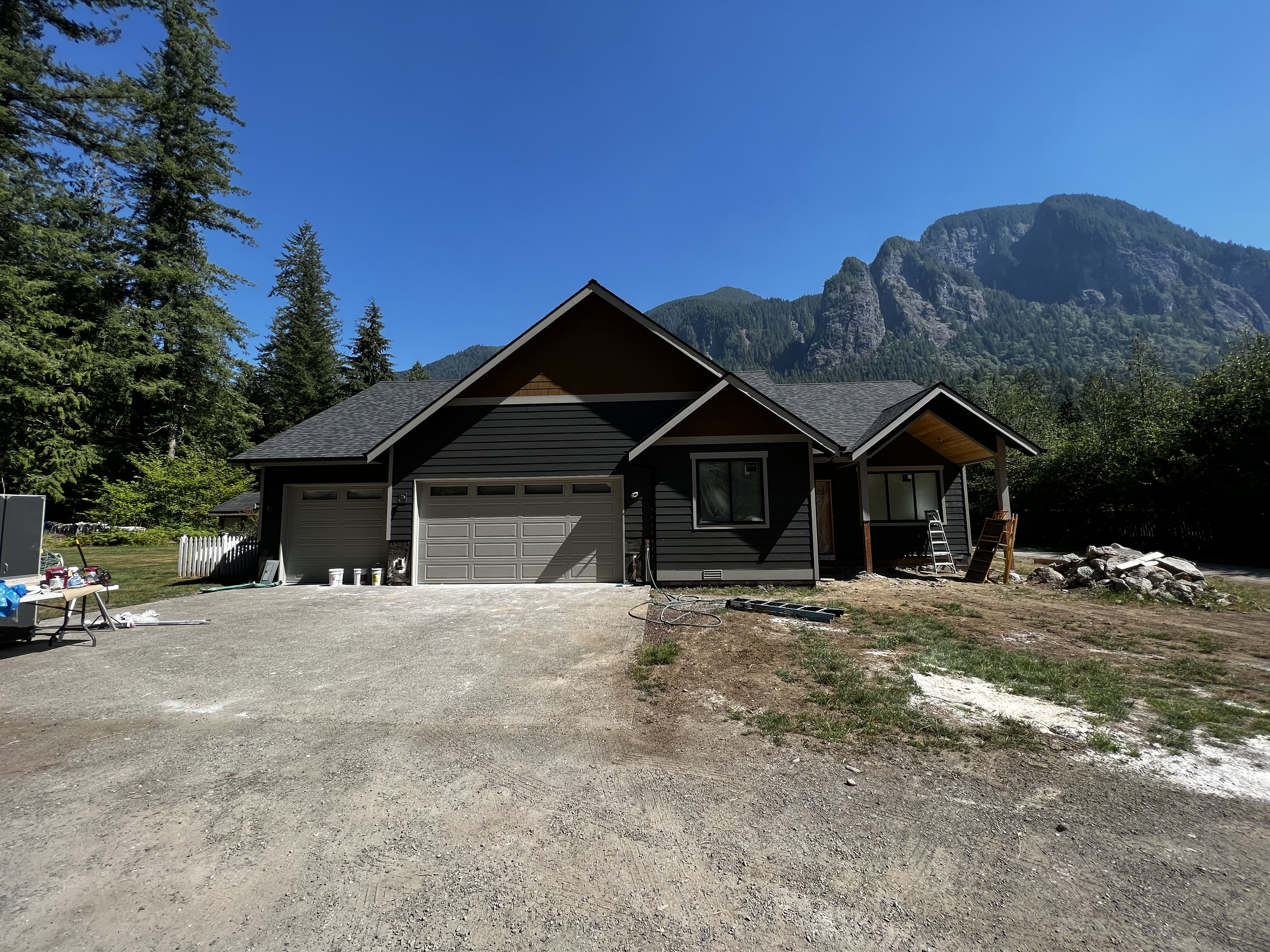 A house under construction with a black exterior, two-car garage, and a front porch, surrounded by trees and mountains in the background, on a clear sunny day.