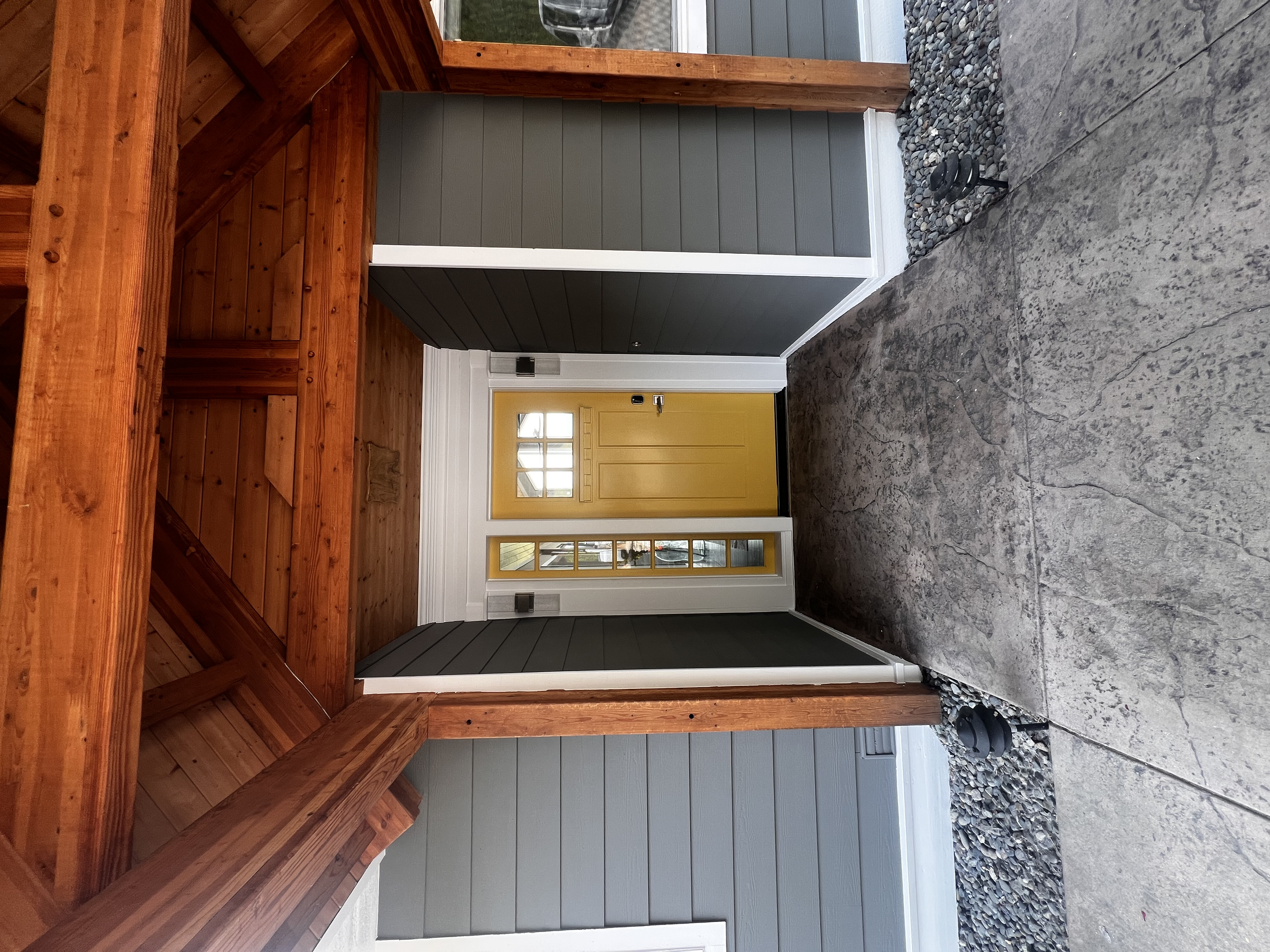 Interior view of a porch area with a yellow front door. The porch has a wooden overhang, gray siding on the walls, a concrete floor, and small rocks lining the edges.