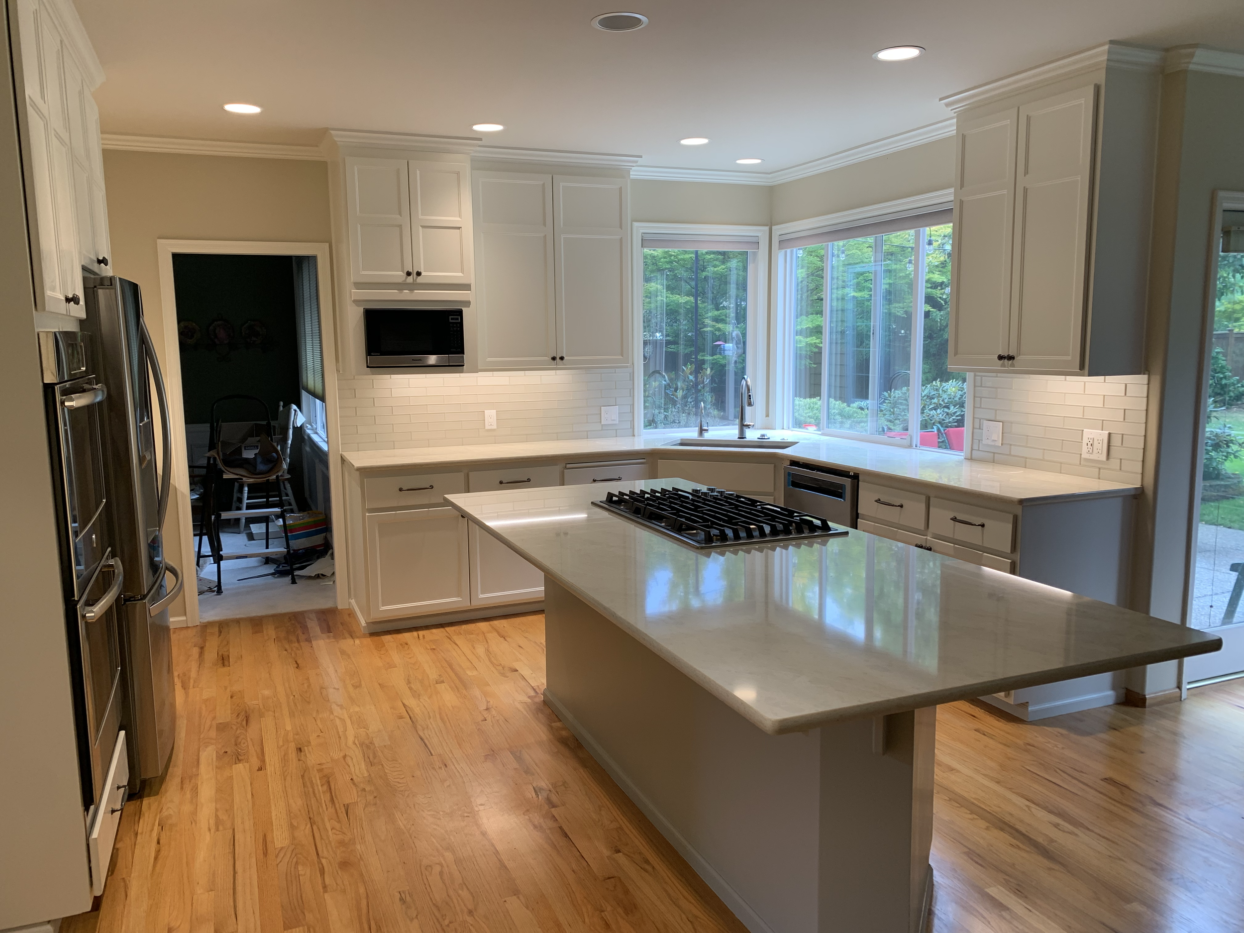 Modern kitchen with white cabinets, granite countertops, a central island with a gas stove, and large windows overlooking a green backyard.
