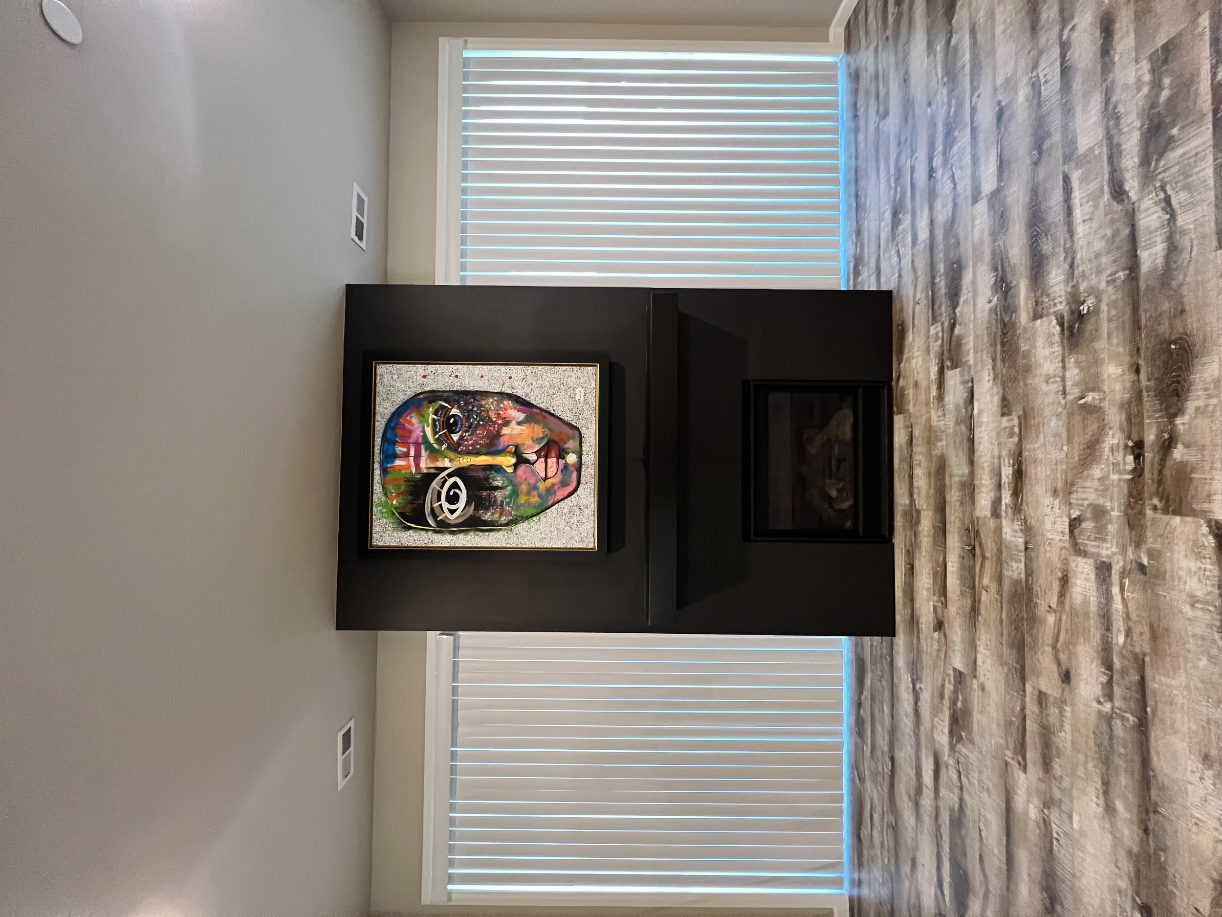 A room with a black fireplace and colorful, abstract artwork of a face hanging above it, with vertical blinds on large windows behind.