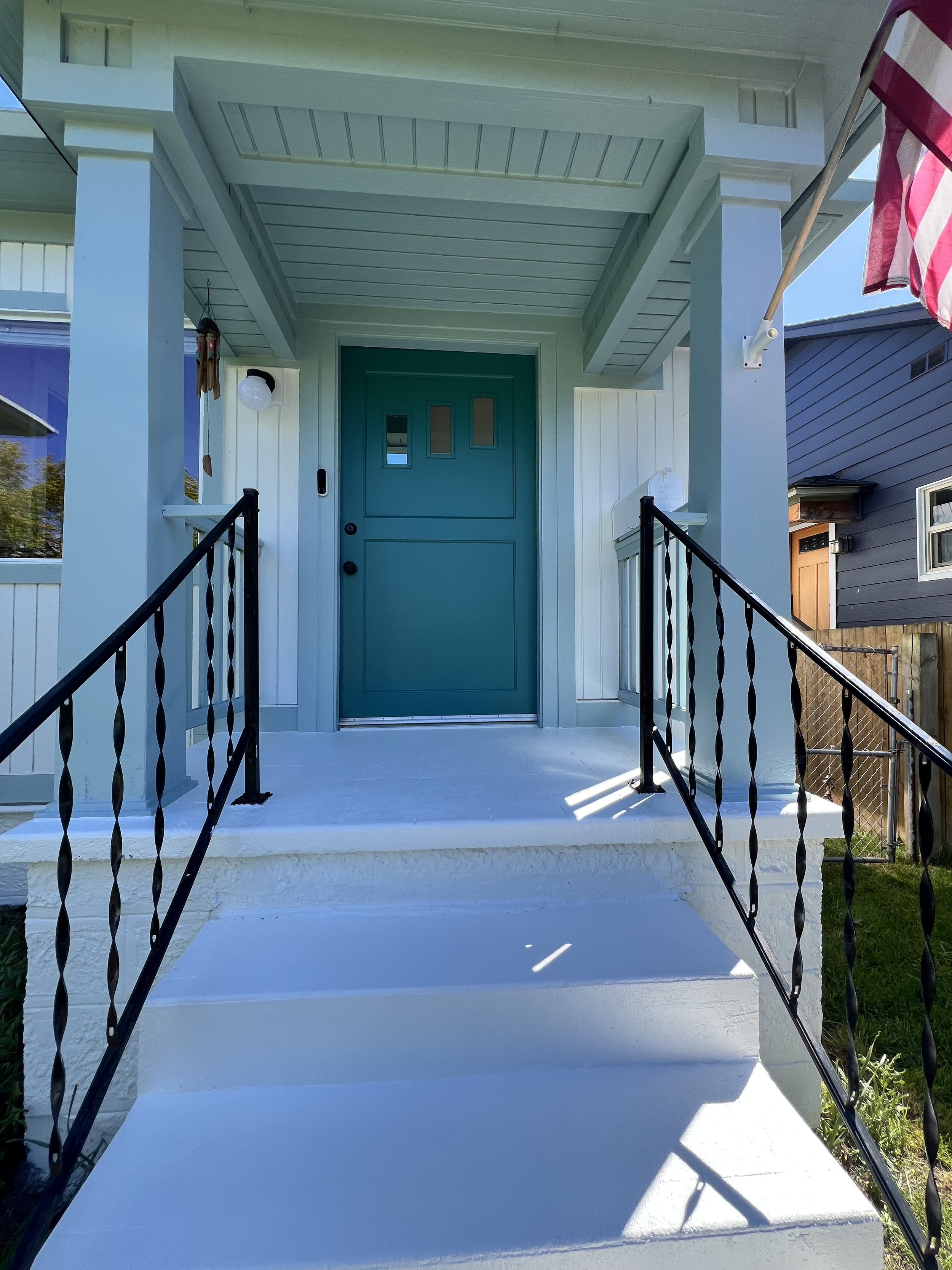 Front porch of a house with a teal door, white siding, black metal railing.