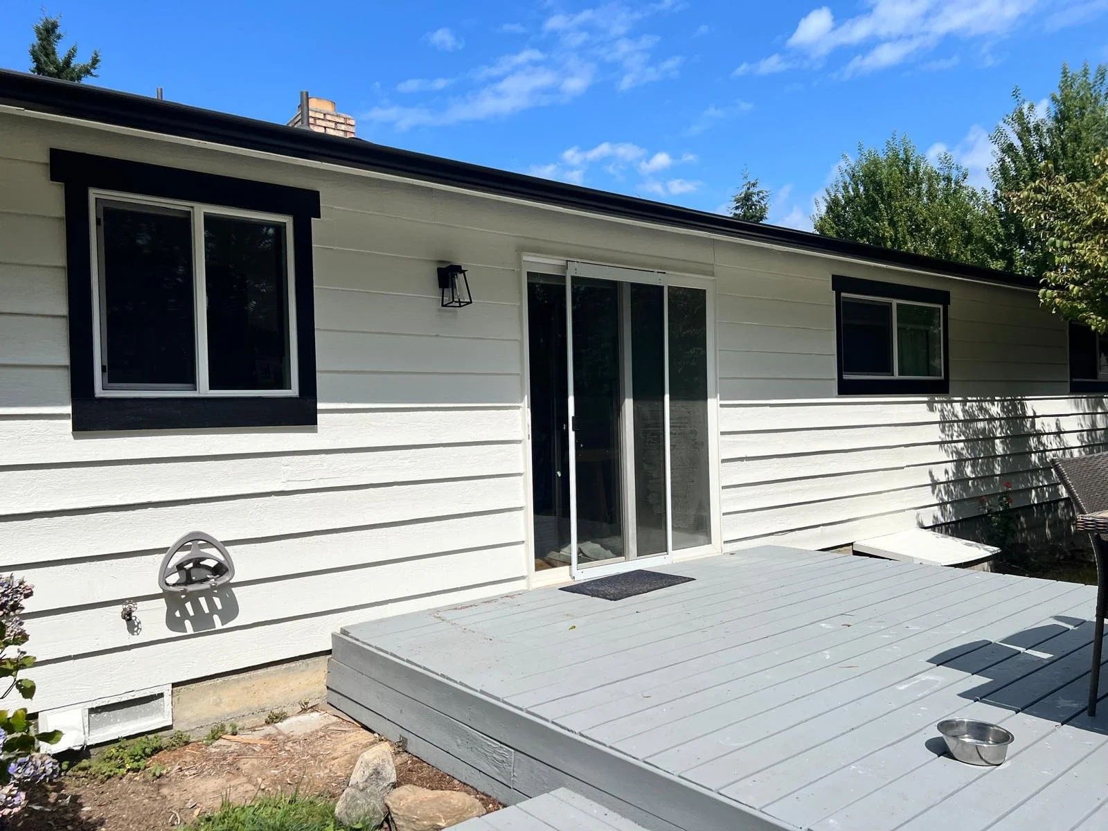 Backyard view of a white house with black trim, featuring a sliding glass door, three windows, a gray deck, and outdoor furniture, under a clear blue sky.