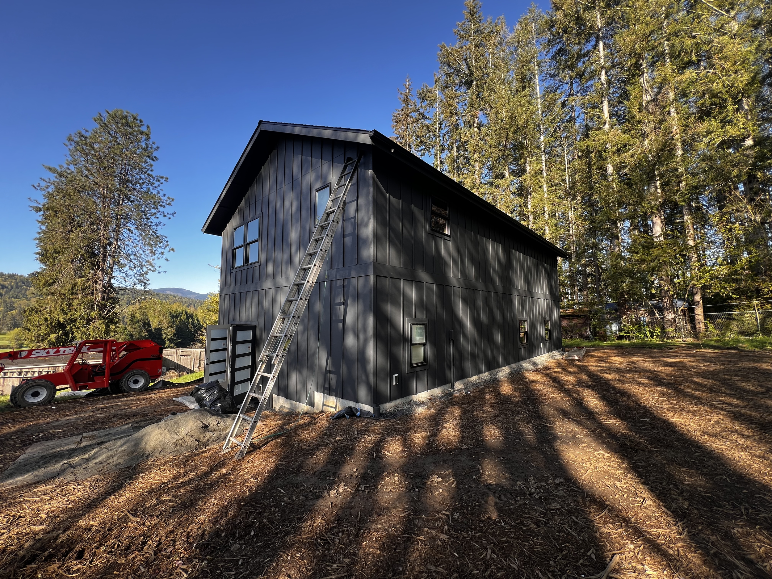 A black metal house under construction in a wooded area with tall trees. A ladder leans against the house, and there is construction equipment nearby, including a red trailer. The ground is covered with dirt and some shadow patterns from the trees.
