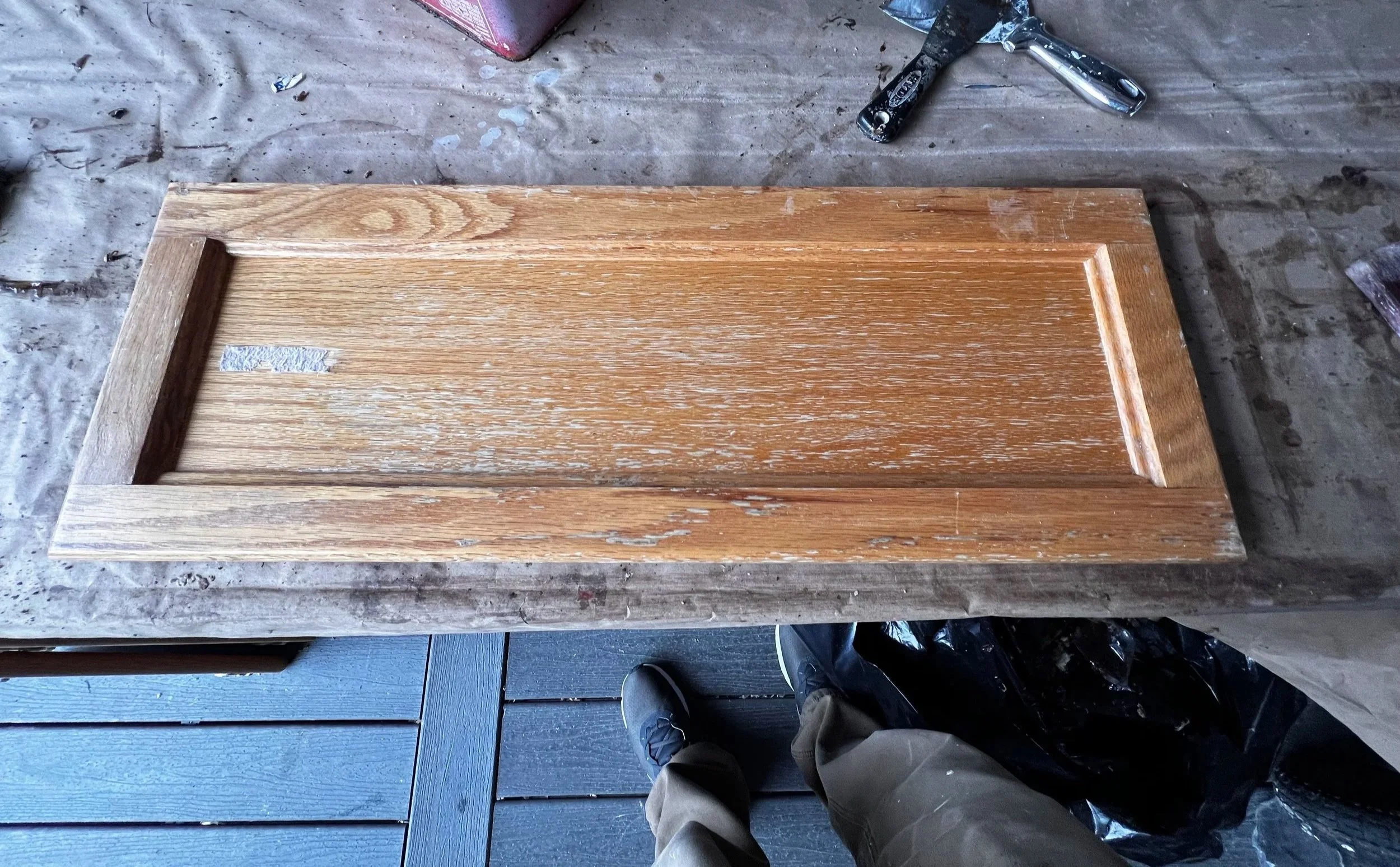 Wooden drawer front on a workbench with tools and a person’s feet visible in the background.