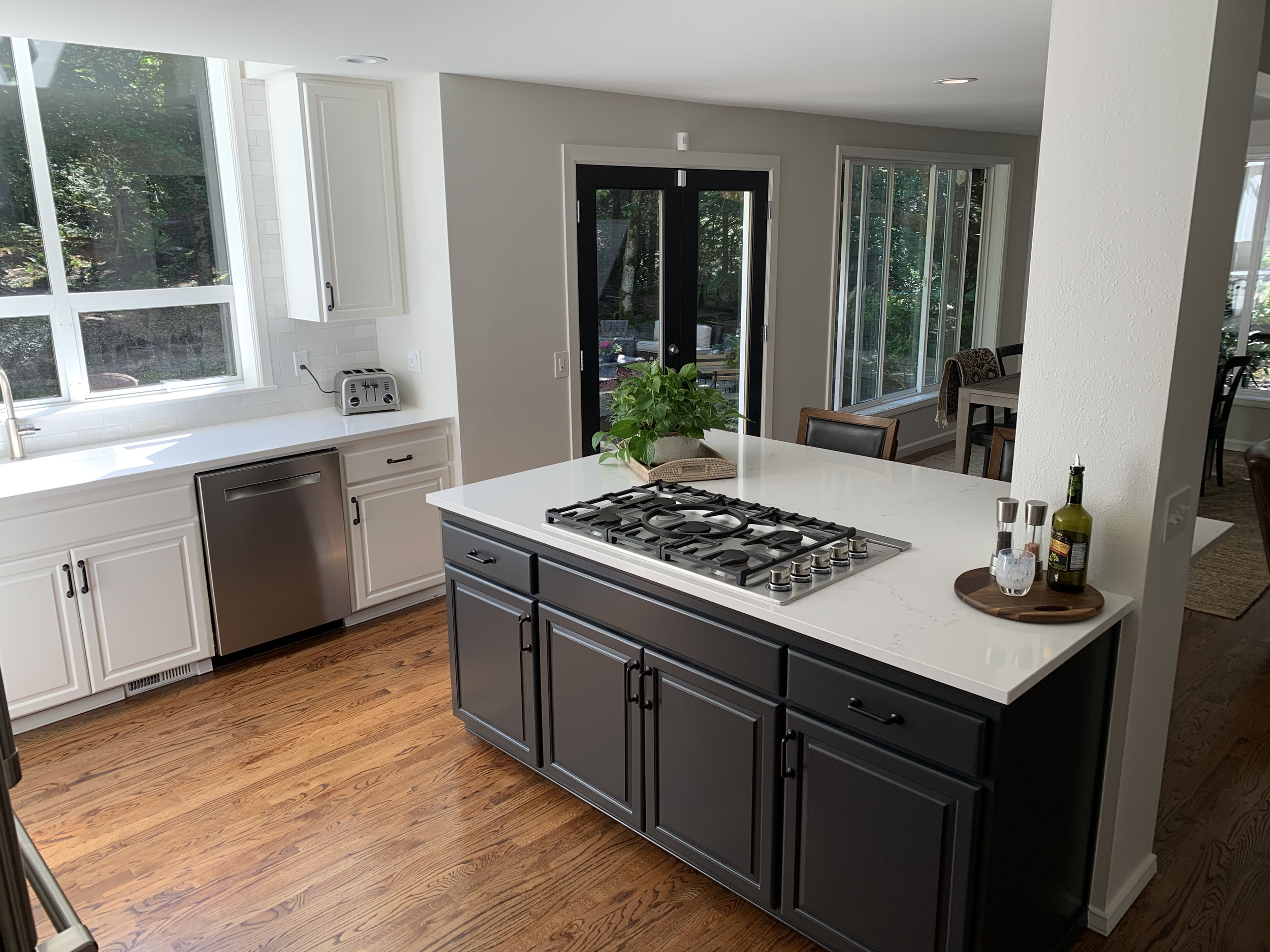 Modern kitchen with white and black cabinets, a white countertop, and wood flooring. There is a gas stove on the island counter, a plant, and some bottles and glasses on a tray. A large window and glass doors provide natural light.