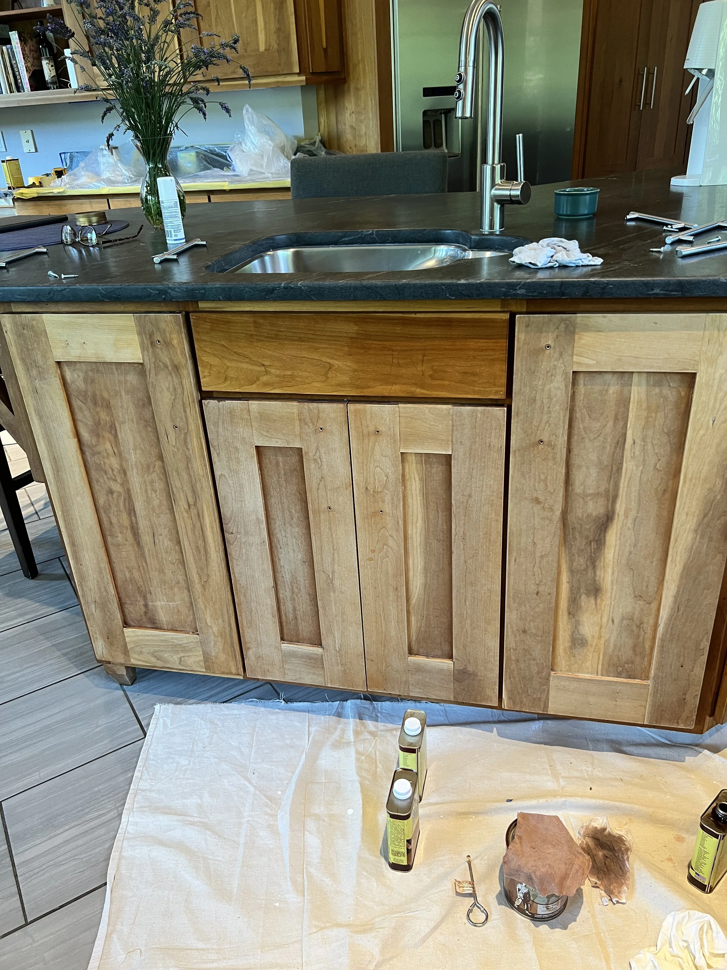 A kitchen with a wooden cabinet and a black countertop. There is a single-basin stainless steel sink with a high arc faucet. On the counter are some tools, a vase with purple flowers, and some paper towels. The floor in front is covered with a white cloth and has painting supplies and brushes.