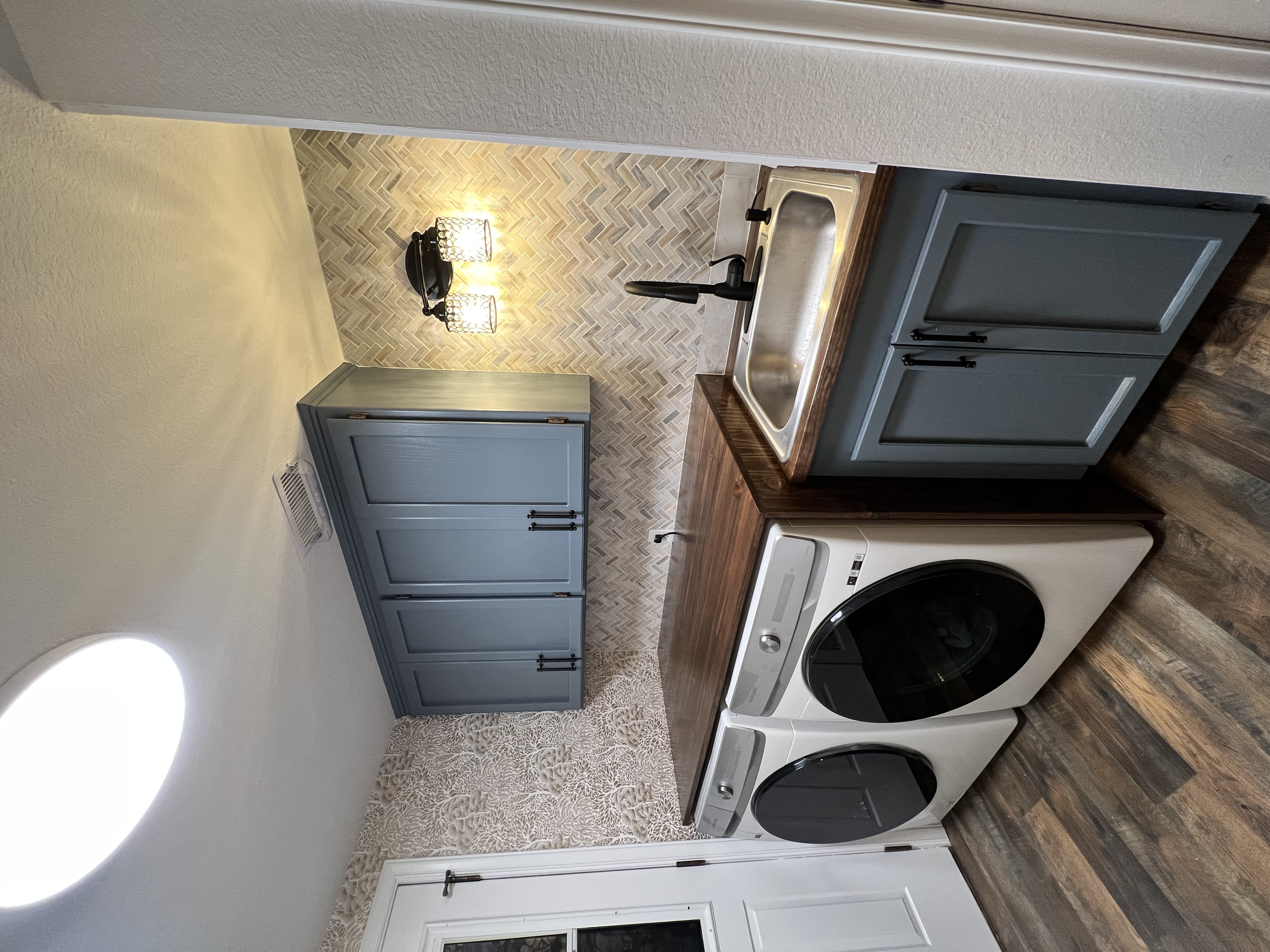 A small kitchen area with a wood countertop, dark gray cabinets, a stainless steel sink with black faucet, a beige chevron tile backsplash, a washing machine, the wall with textured wallpaper, and a ceiling light.