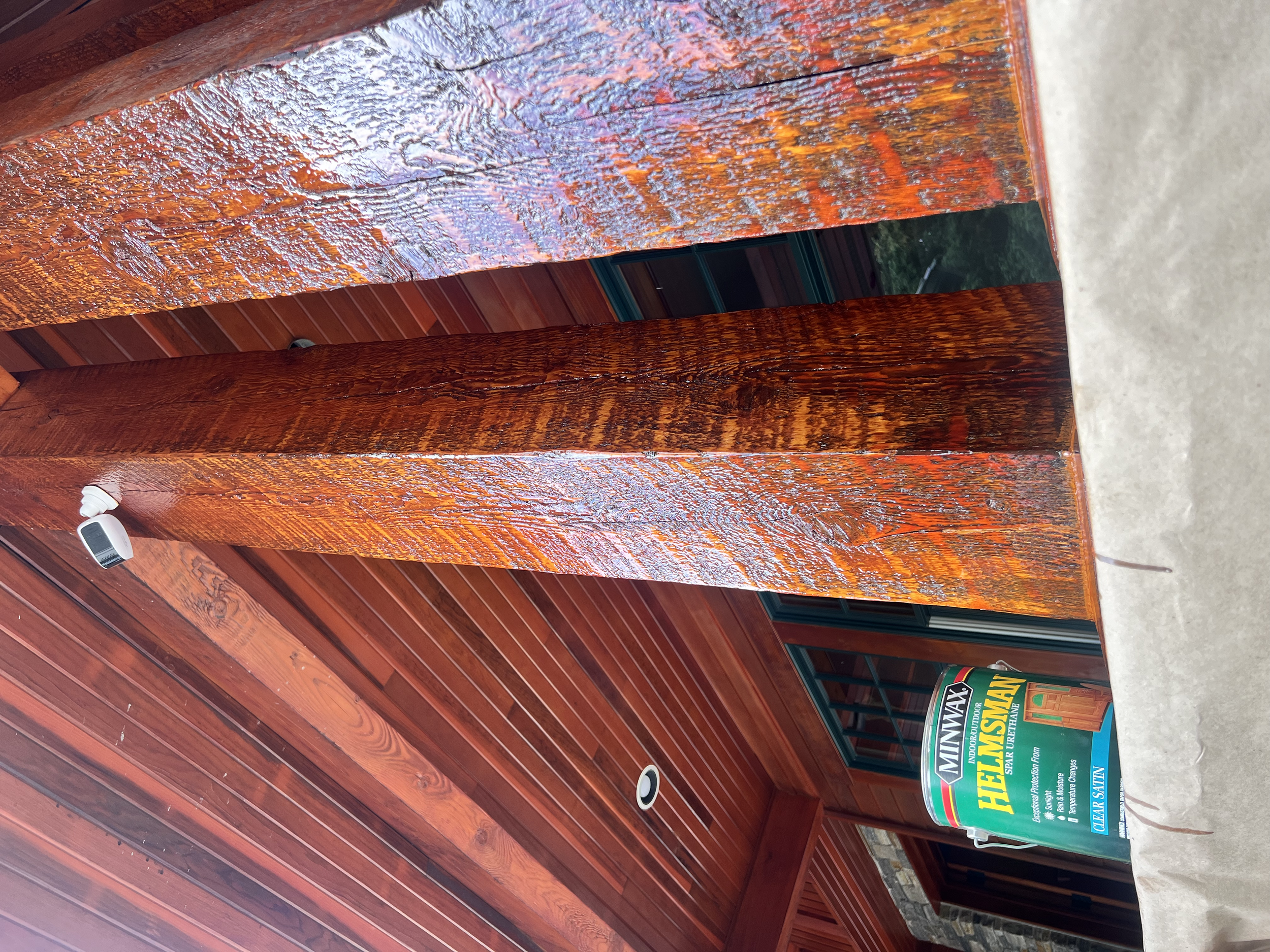 Close-up of a wooden deck with a dark green window frame in the background. A Minwax Helmsman polyurethane sign is partially visible on the right side. The wood has a glossy finish, showing the wood grain and texture.