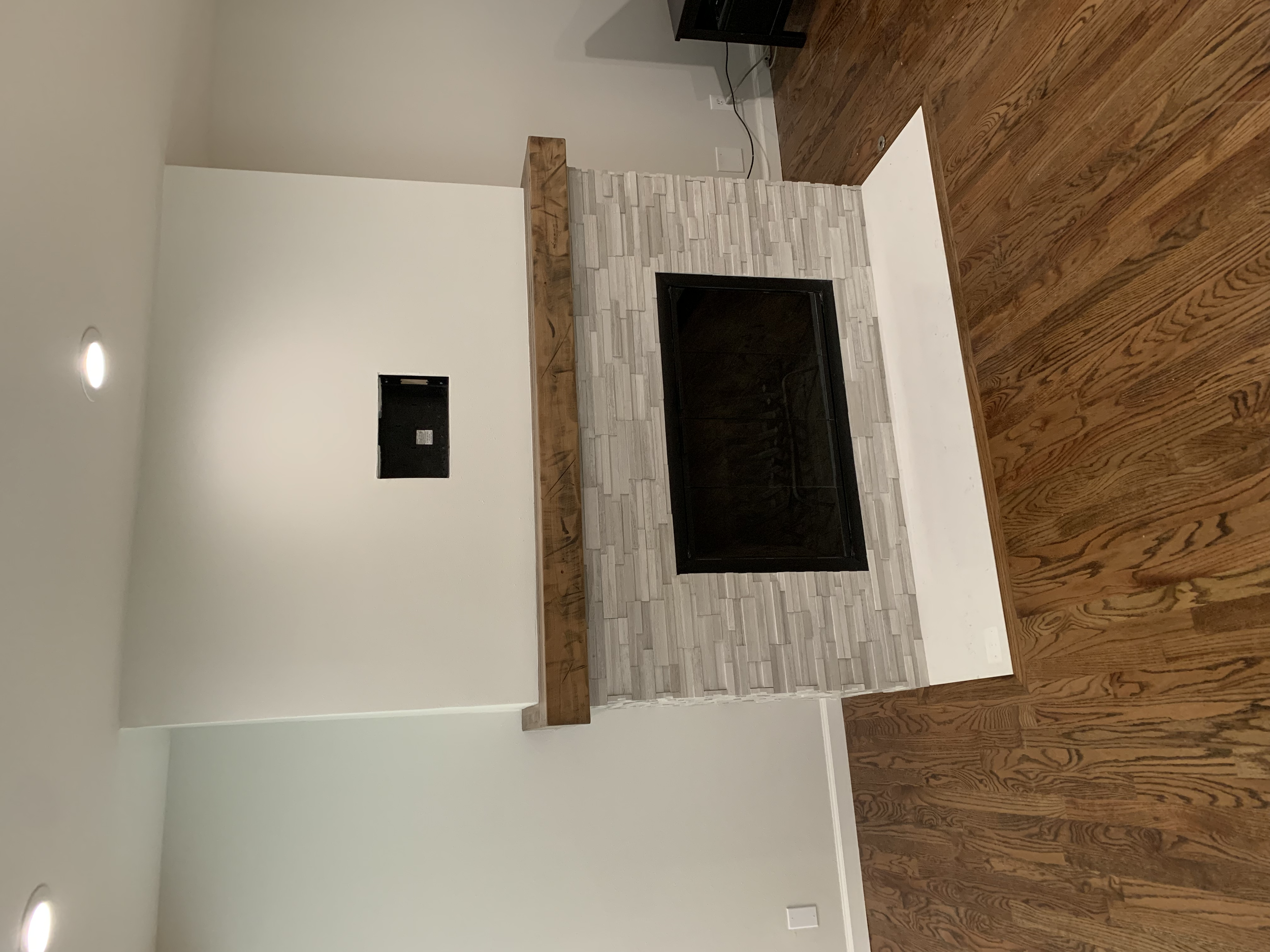 A fireplace with a gray tiled surround and a black metal frame, topped with a wooden mantel, in a room with white walls and hardwood flooring.