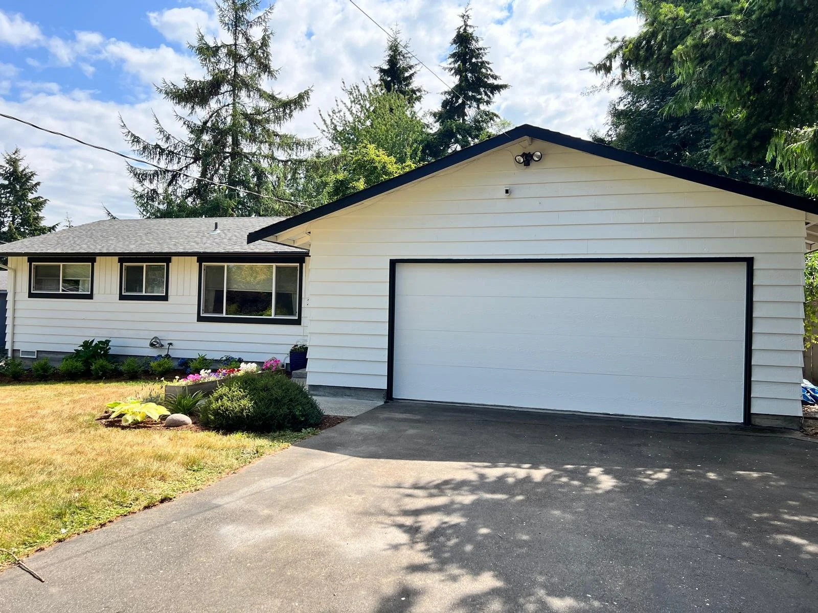 Front view of a white house with black trim, a large garage door, a driveway, and a garden with flowers and shrubs. Tall trees and a partly cloudy sky are in the background.