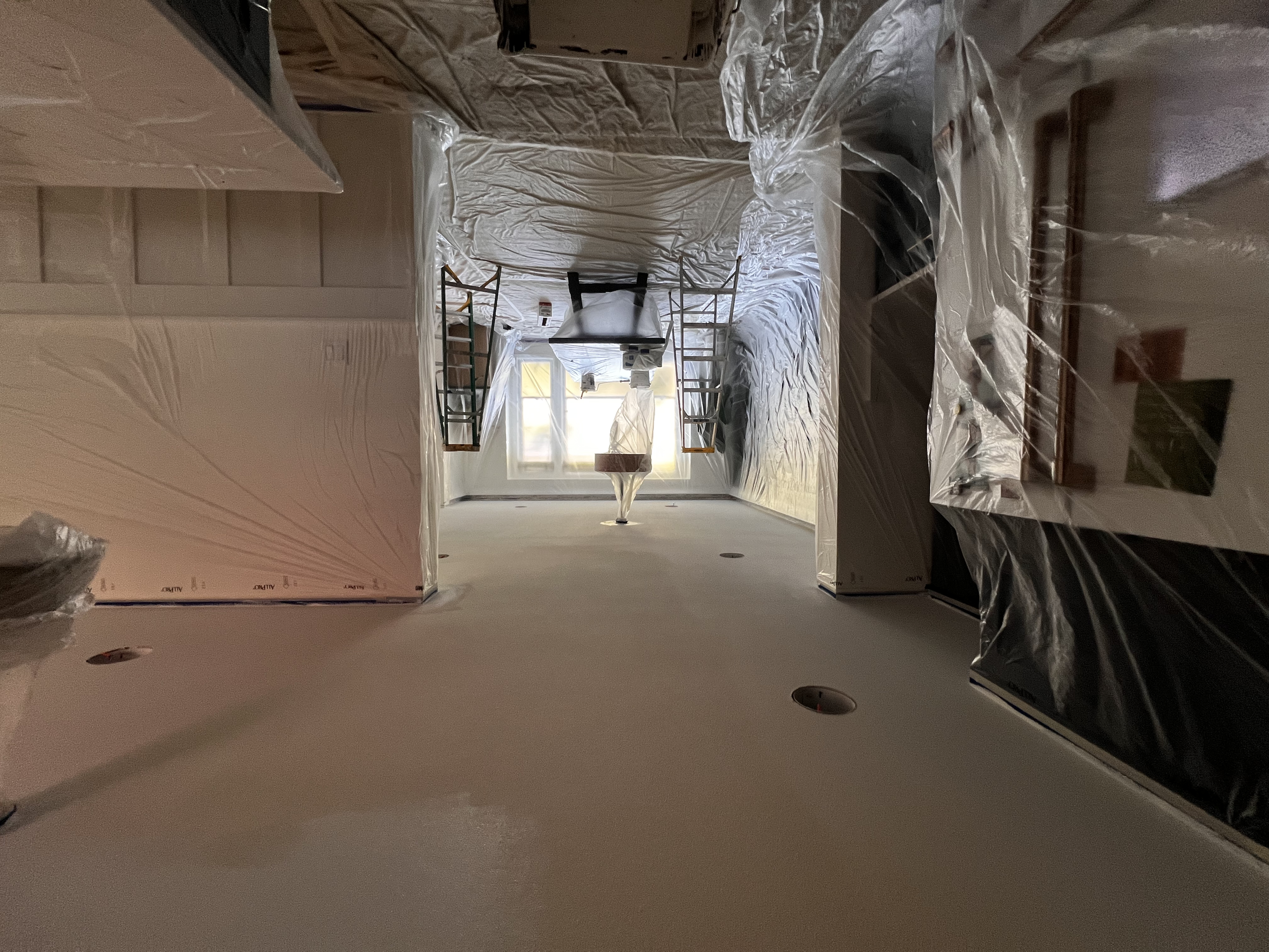 A room under renovation with plastic sheeting covering the ceiling and walls, and construction tools and furniture, including ladders and a table, seen near a window.