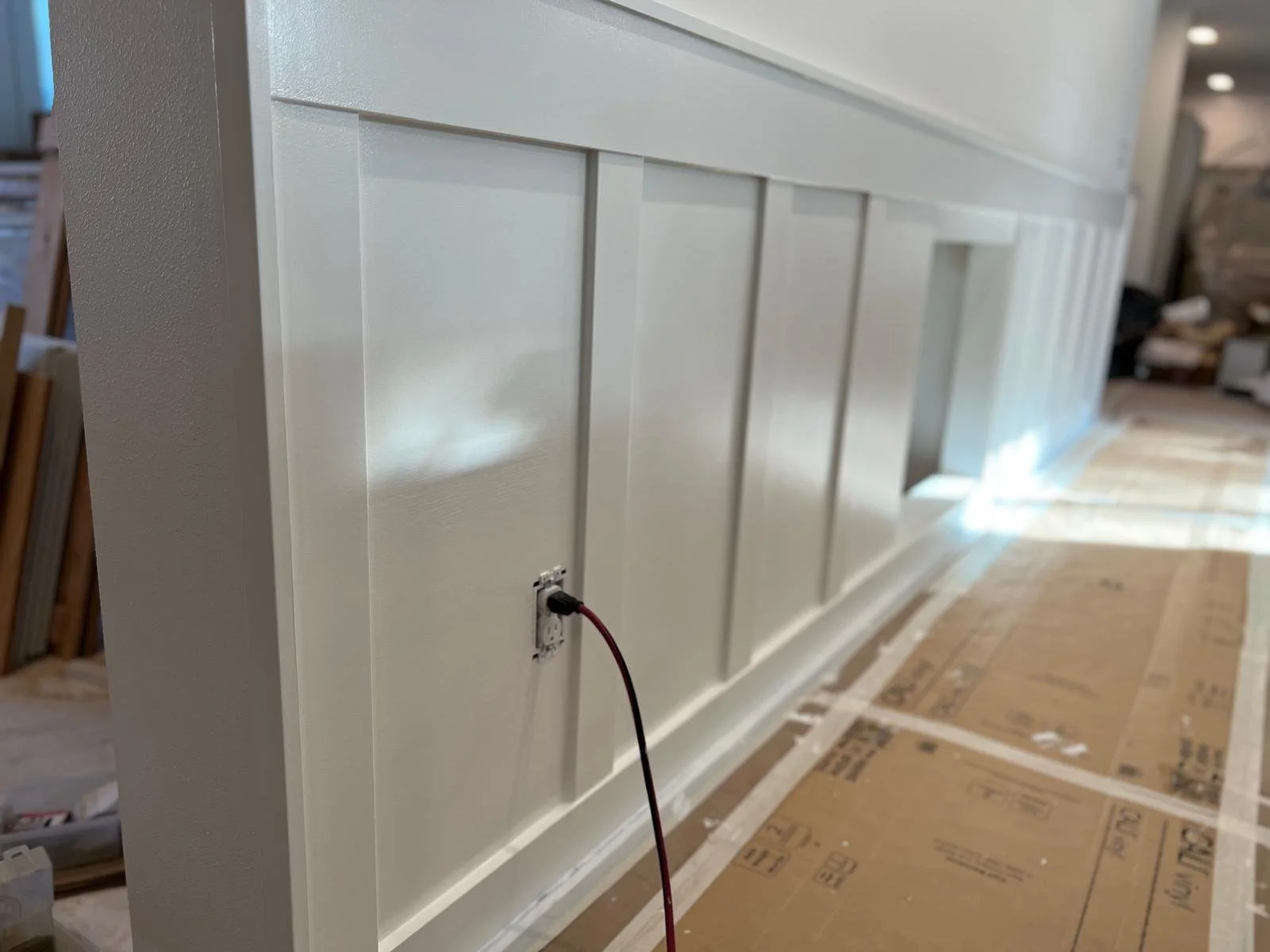 White paneled wall with an electrical outlet and power cord, under construction in a room with cardboard-covered floor.