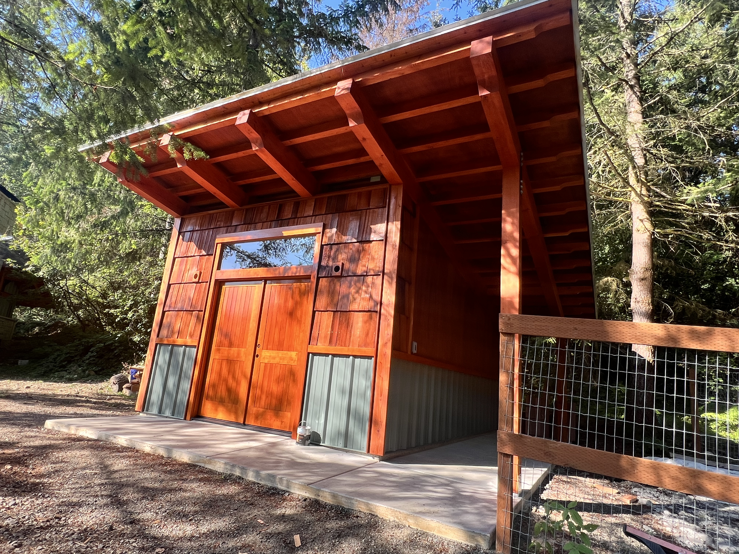 A small wooden shed with a metal base, surrounded by forest trees. The shed has a sliding door, a window above, and a sloped roof, with sunlight casting shadows from nearby trees.