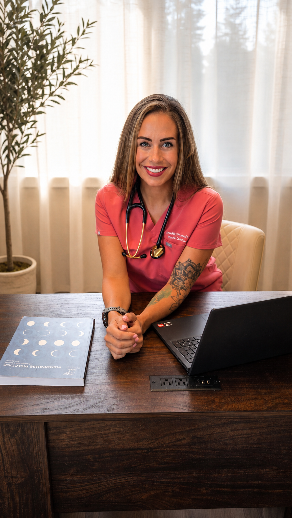 A smiling woman in pink scrubs sitting at a wooden desk with a laptop and a book, with a stethoscope around her neck and tattoos on her left arm.