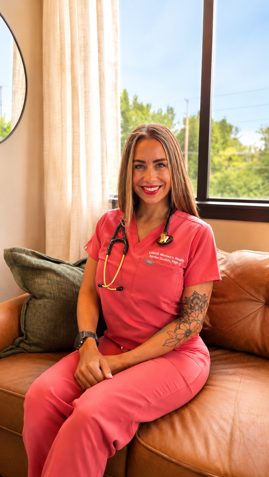 A female healthcare professional in pink scrubs sitting on a leather couch, smiling, with a stethoscope around her neck, inside a room with a large window and beige curtains.