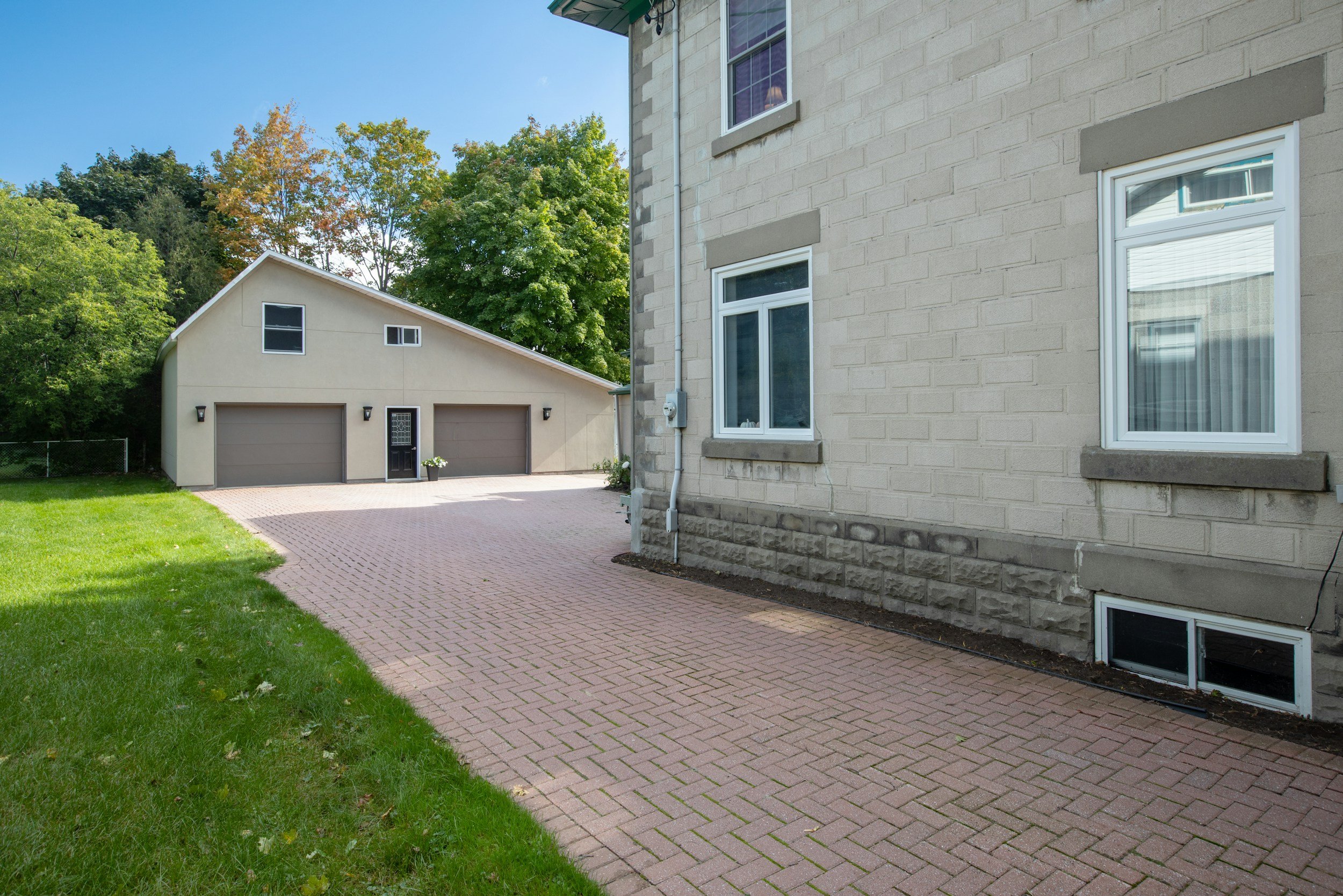 View of a residential backyard with a brick driveway, a garage with two doors, a beige stone house with white-framed windows, a fence, and green trees under a clear blue sky.