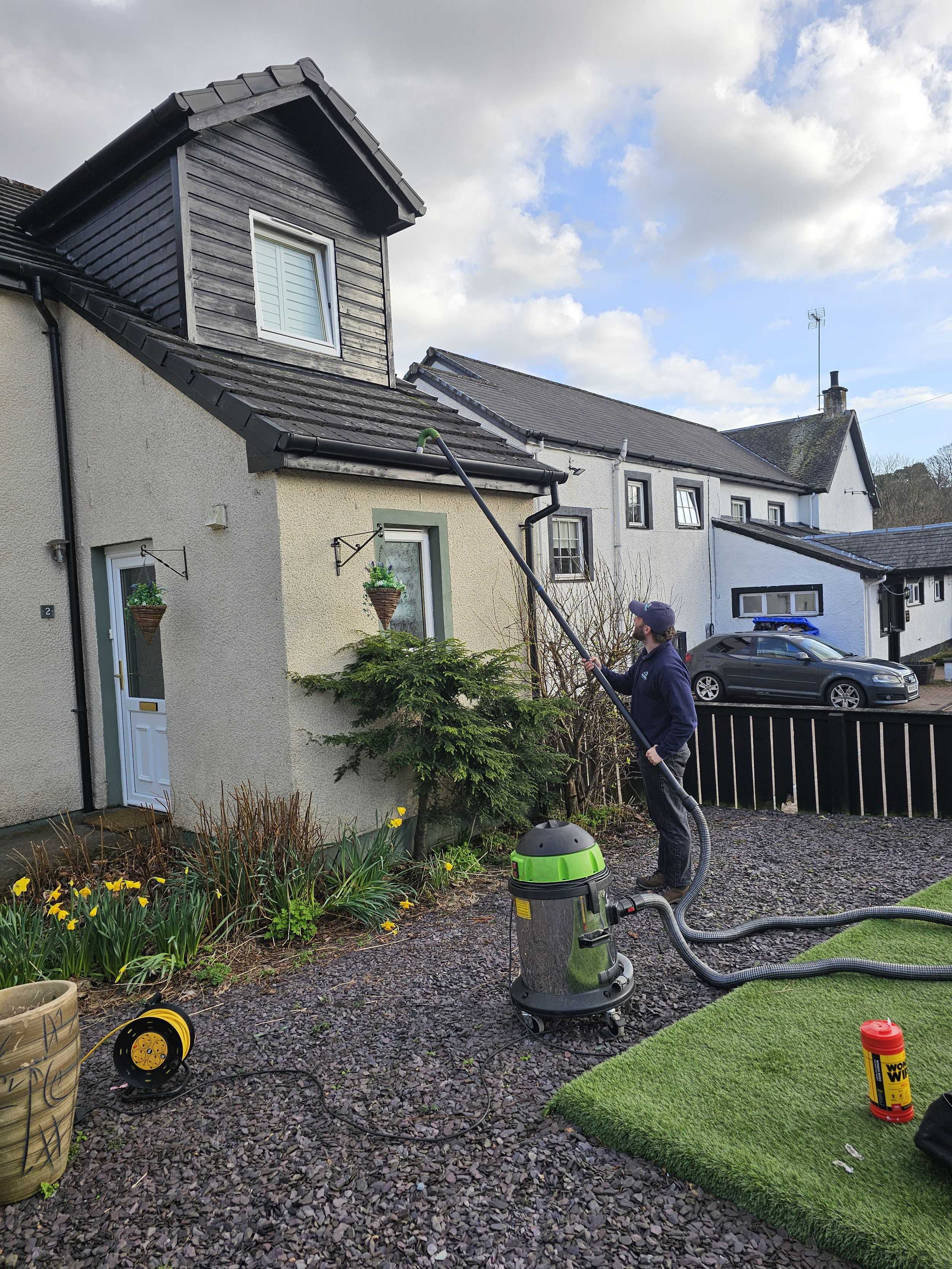 A person using a wet/dry shop vacuum cleaner outside a house, cleaning the garden area with a hose attachment. The house has multiple windows, hanging flower baskets, and a black fence. There are gardening tools, a yellow extension cord, and a patch of artificial grass on the gravel ground.