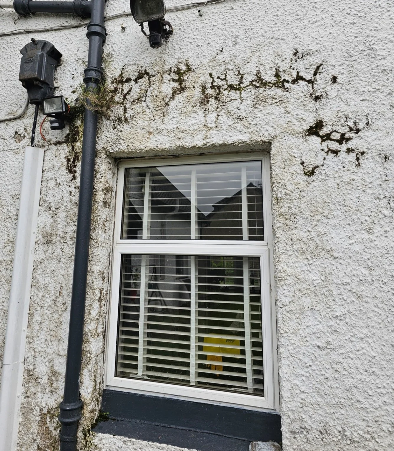 Exterior view of a house wall with a window, drainpipes, and electrical components. The wall surface is textured and shows moss and cracks.
