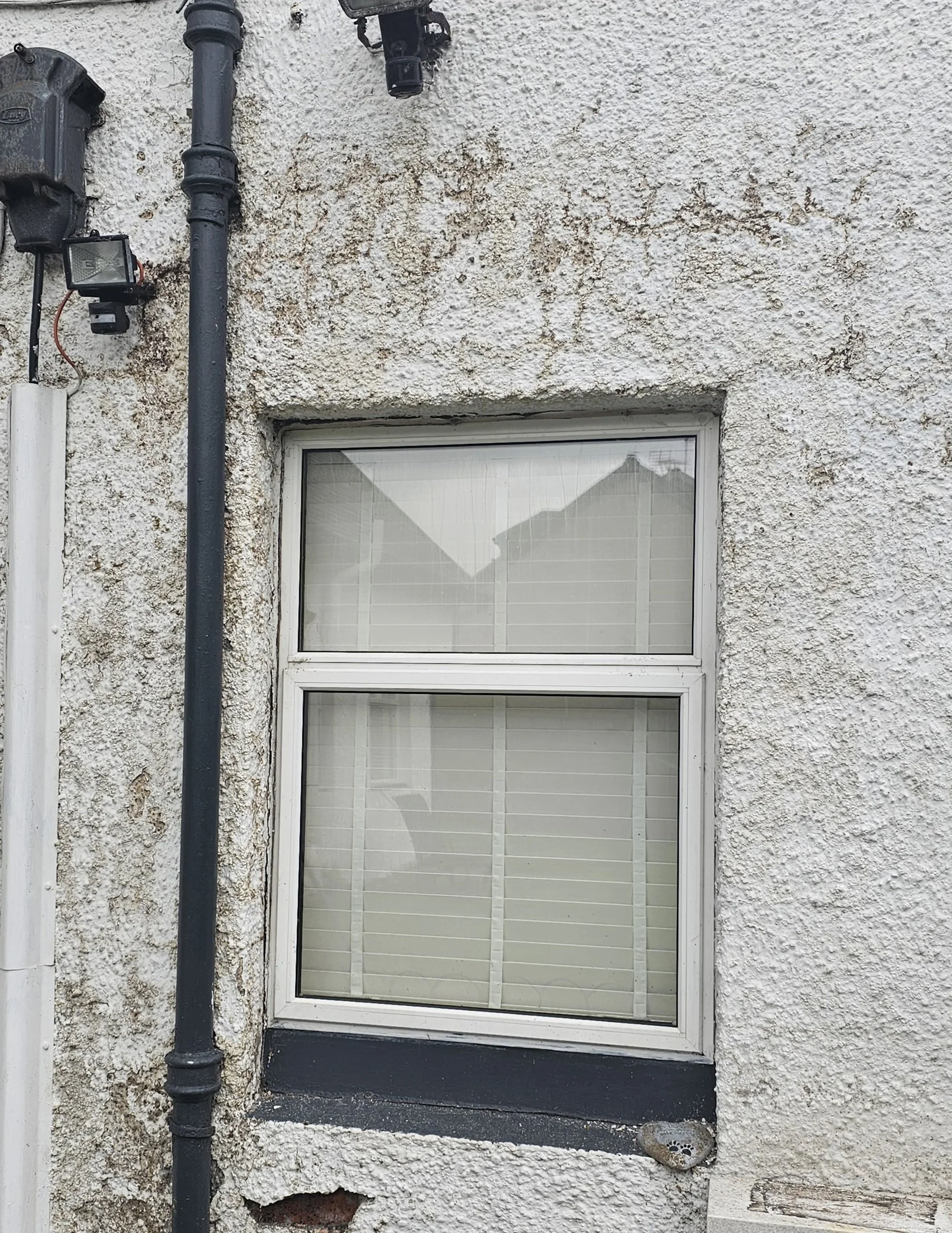 Exterior wall of a building with a window, a black pipe, a small outdoor light fixture, and a rock on the ground with a circular pattern.