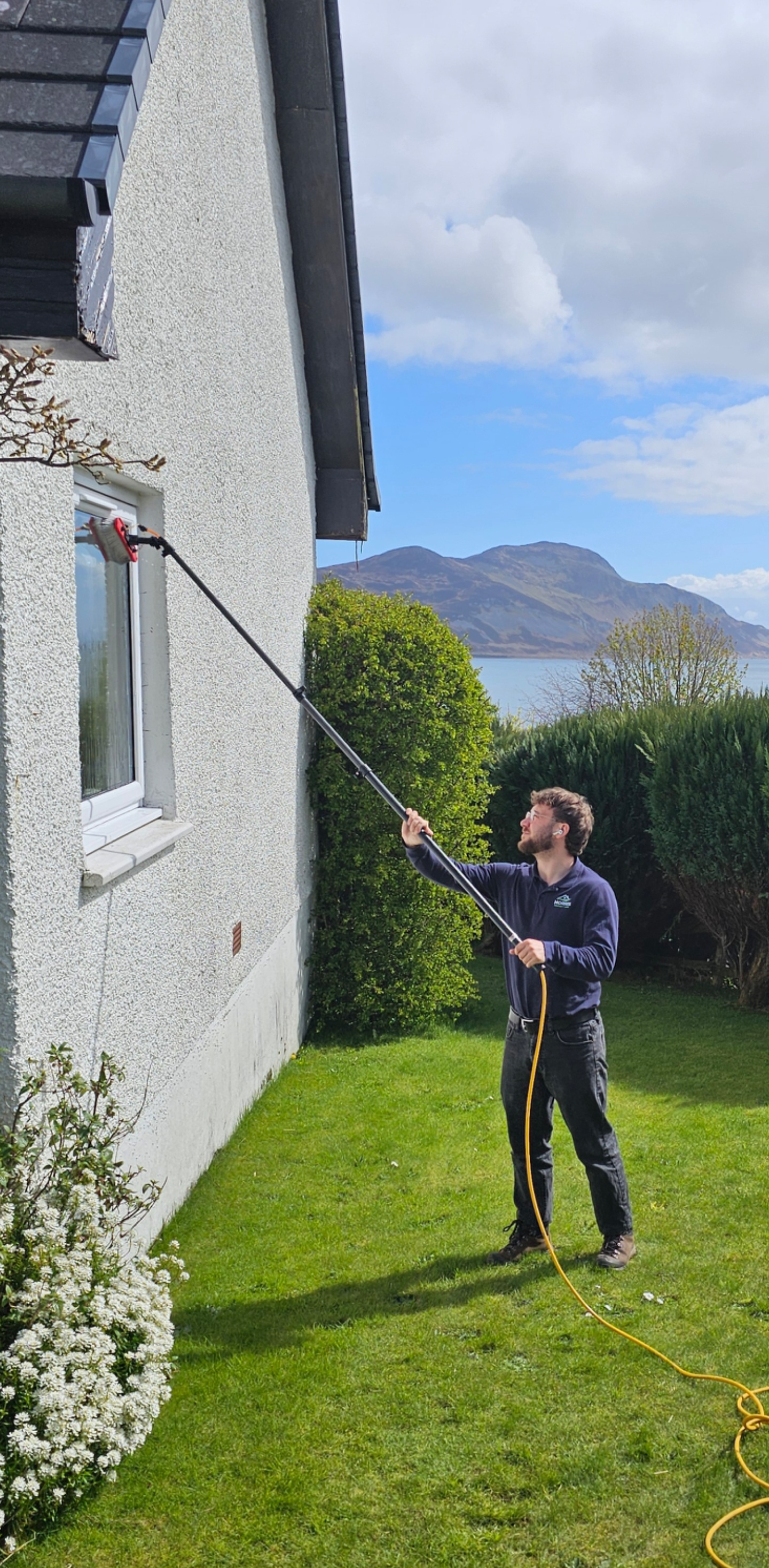 A man cleaning the exterior window of a house with a long-handled cleaning tool, outdoors on a grassy lawn with bushes, trees, mountains, and a lake in the background.