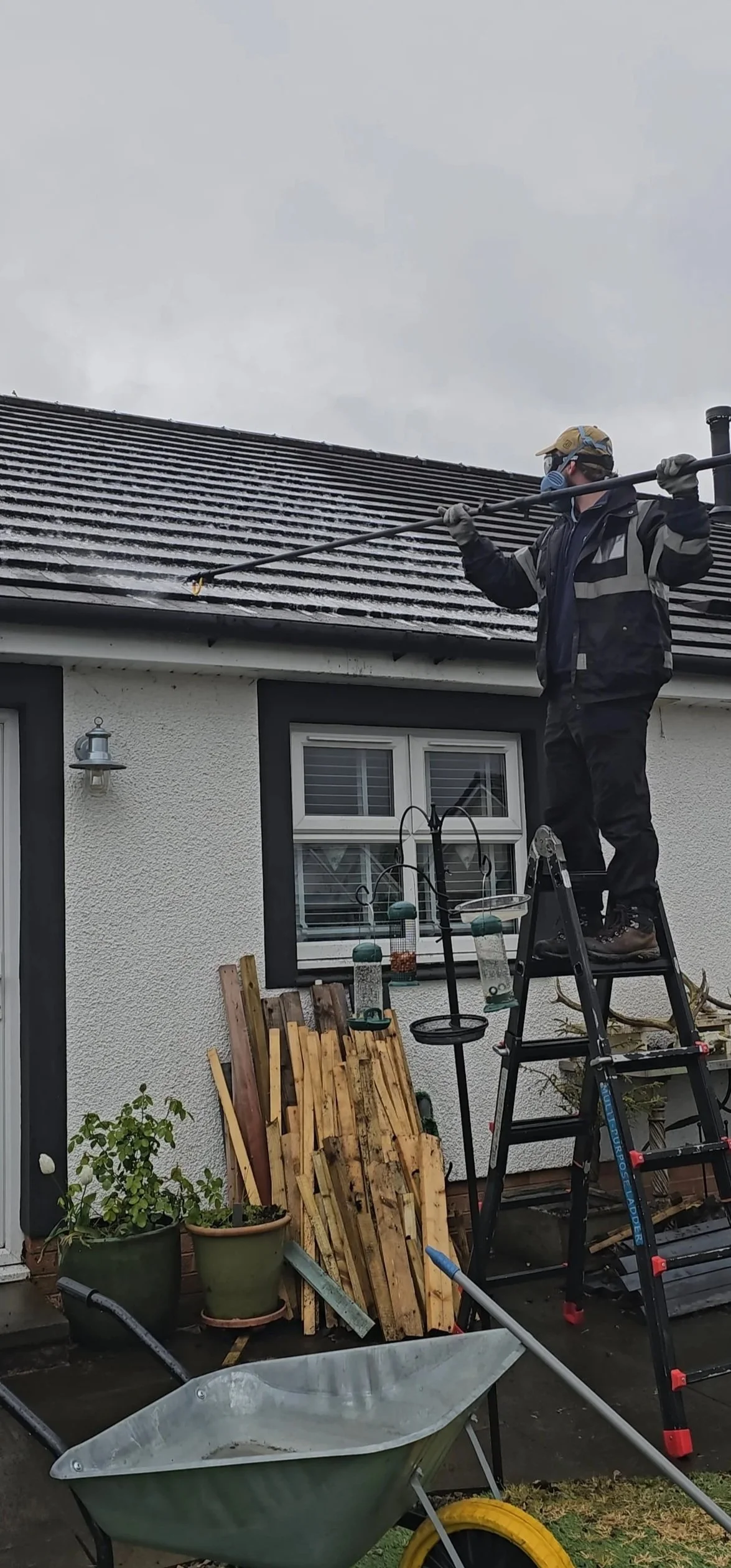 A man wearing a mask, gloves, and outdoor gear is standing on a ladder, using a pressure washer to clean the roof of a house. There are potted plants, wooden planks, bird feeders, and a wheelbarrow in the yard.