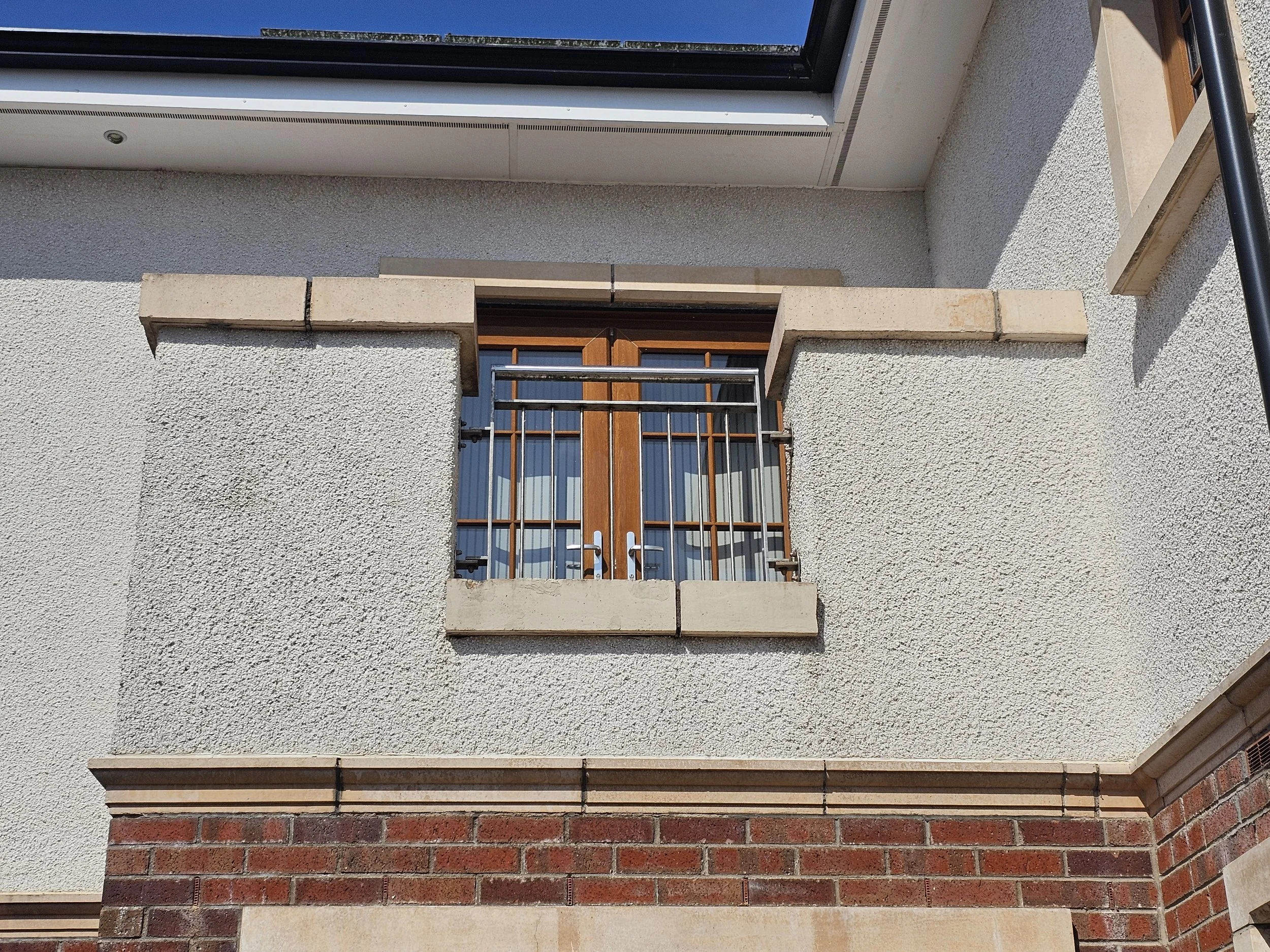 Close-up of a house wall featuring a textured white surface, with three horizontal decorative stone trims, and a lower row of red bricks. A wooden door with a metal security grille and a window are part of the scene.