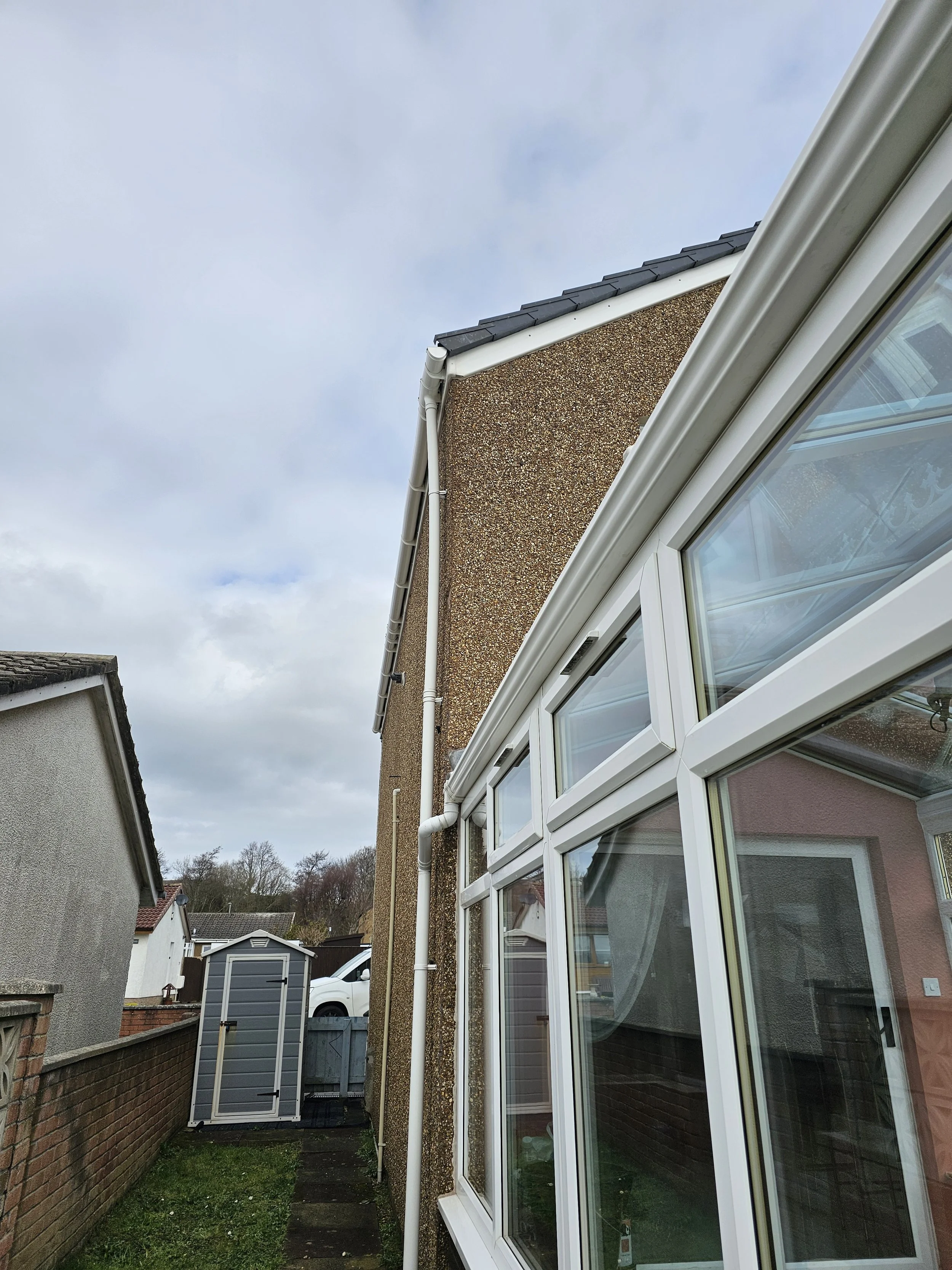 Side view of a building with a peaked roof, white-framed windows, and rain gutter, with a backyard garden and shed in the background under a cloudy sky.