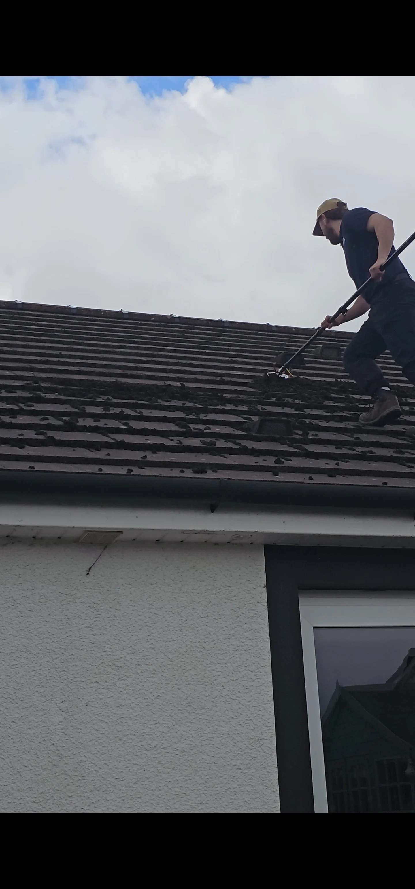 A man wearing a cap and black shirt cleaning or working on a sloped roof with dark shingles, using a long-handled tool, under a partly cloudy sky.
