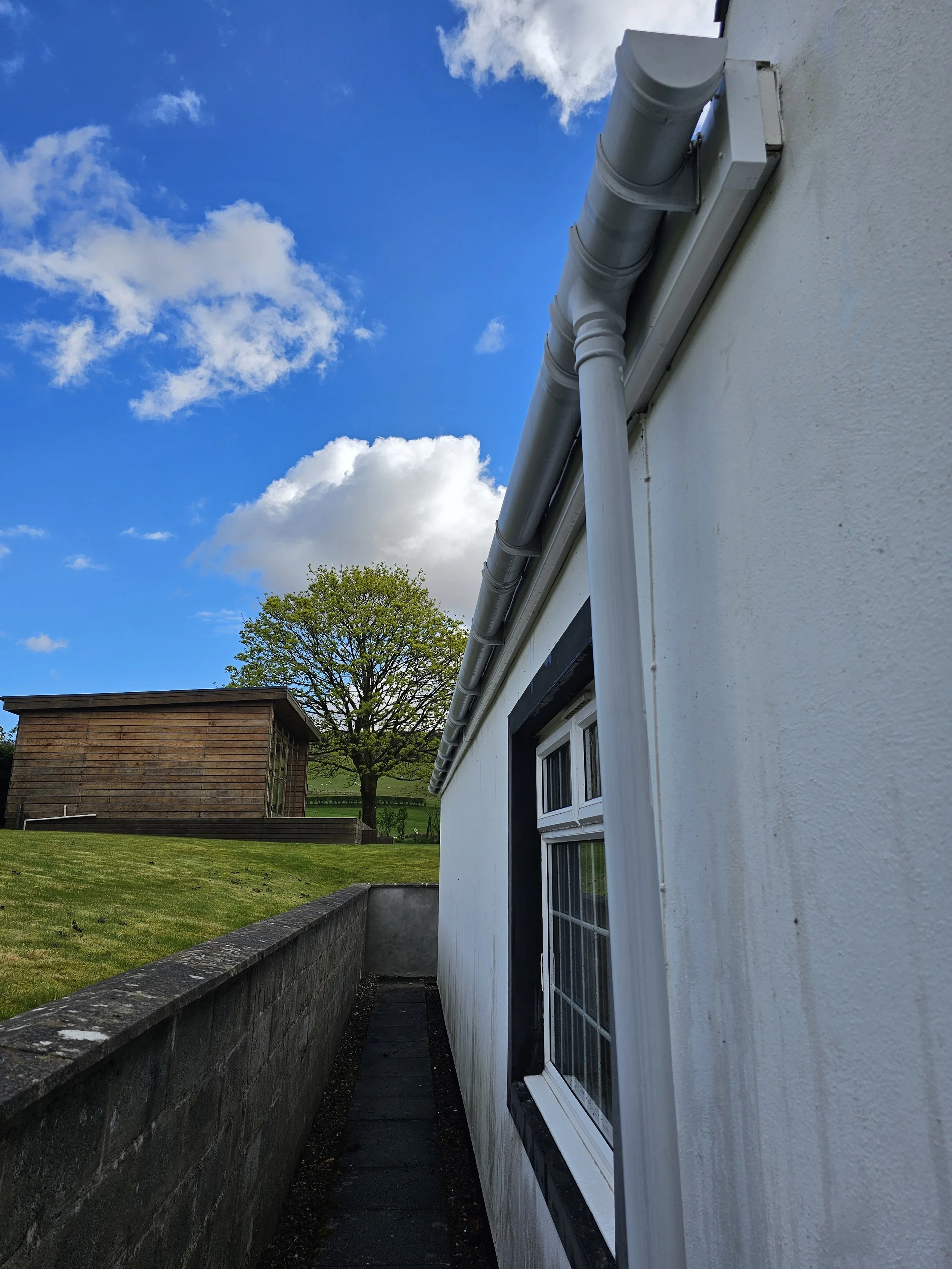 View from the side of a white building showing outdoor gutter system with yellow-green grass, a wooden shed, trees, and partly cloudy blue sky in the background.