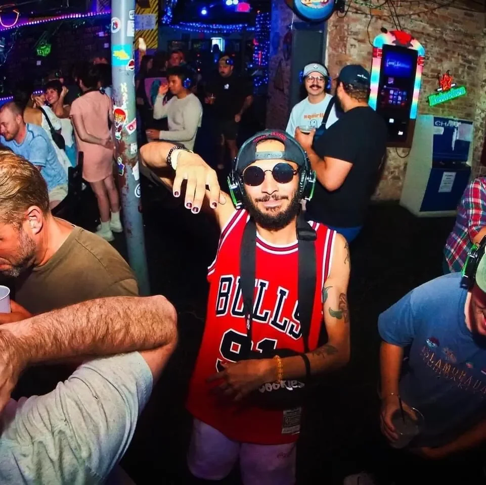 Person wearing sunglasses and a Chicago Bulls basketball jersey at a lively bar or nightclub, with other people in the background enjoying the music and socializing.