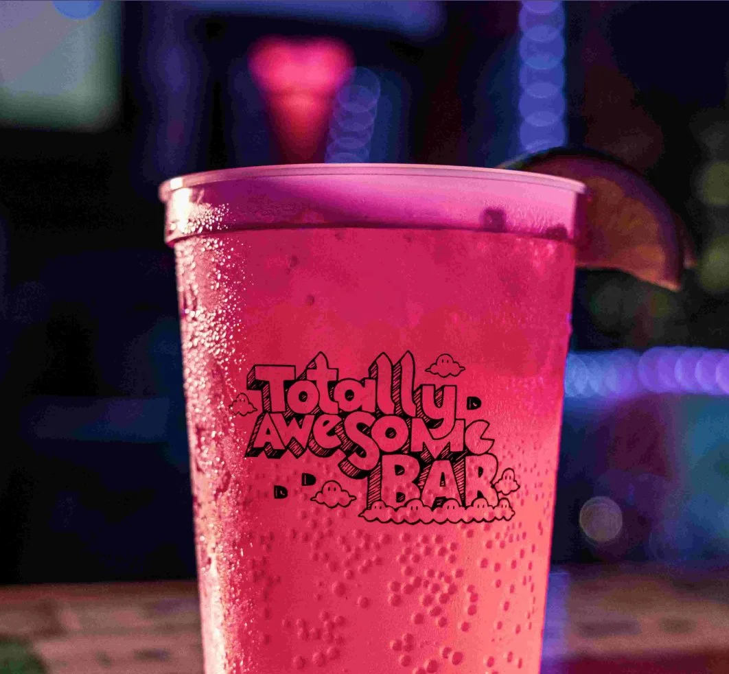Close-up of a pink plastic cup with condensation, displaying the text 'Totally Awesome Bar' with small clouds, surrounded by blurred colorful lights.