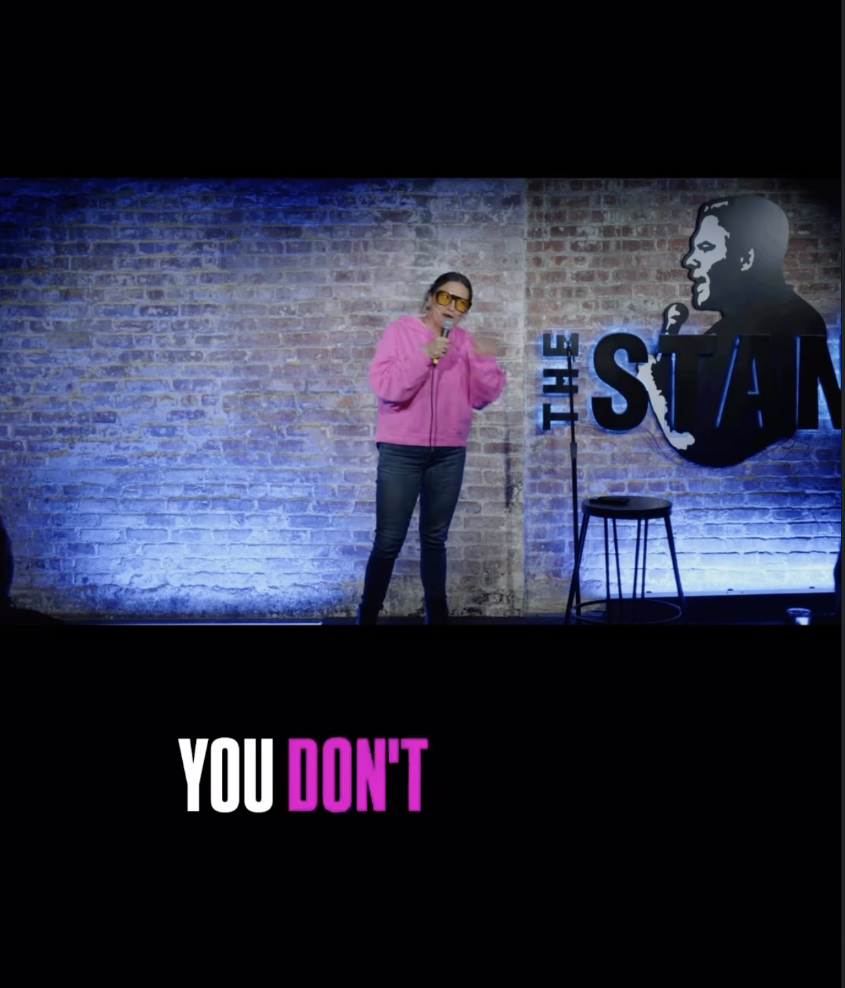 A woman wearing a pink hoodie and sunglasses performing comedy on stage at The Stand comedy club, with a brick wall background and a sign of a man holding a microphone.