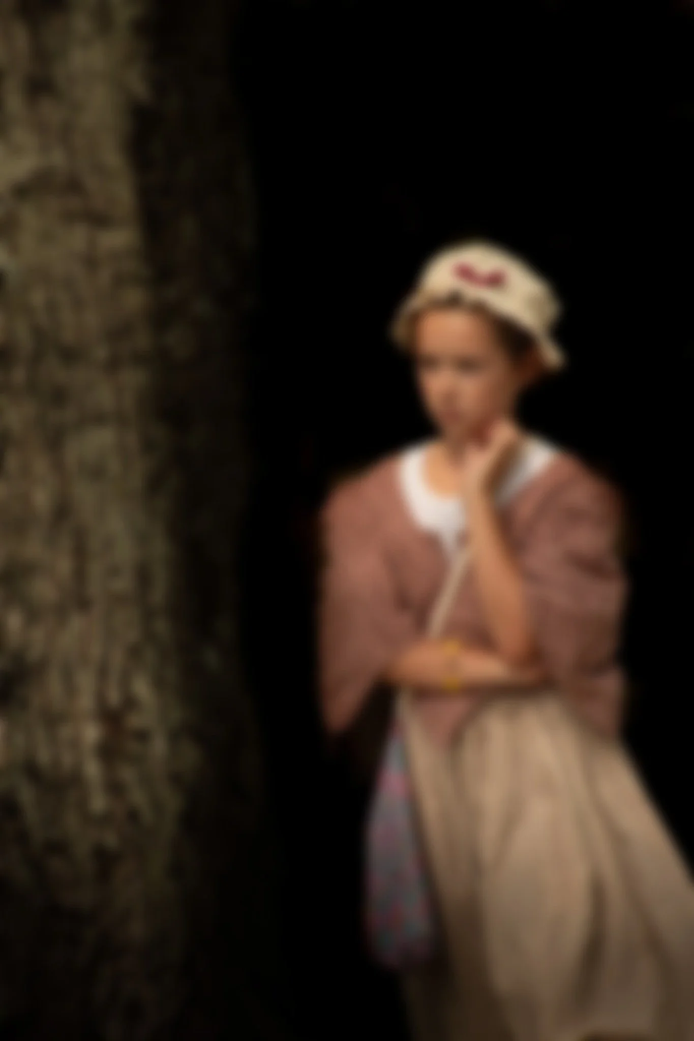 A child wearing a vintage style dress and hat, standing next to a tree with a thoughtful expression and a dark background.