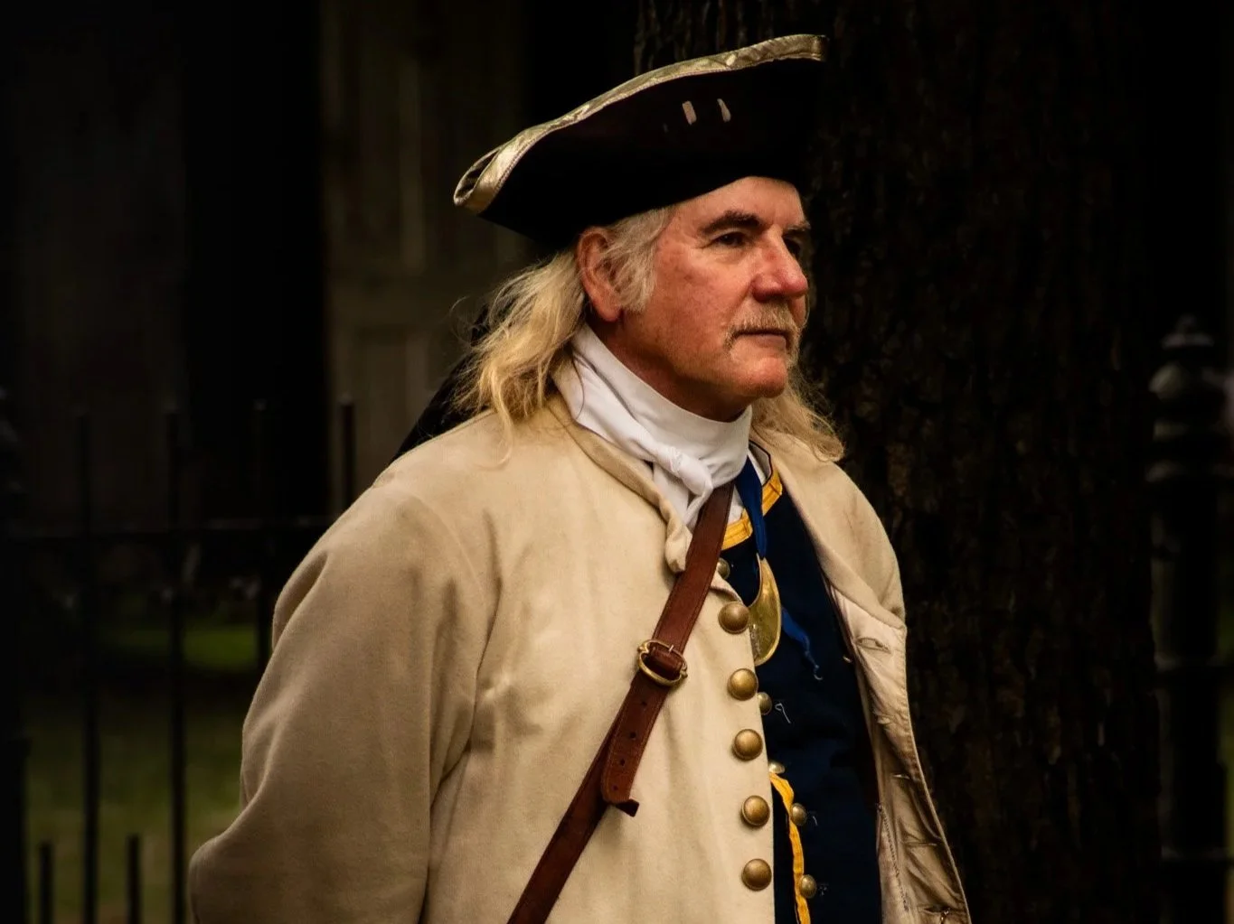 A man dressed as a historical soldier from the American Revolutionary War, wearing a tricorn hat, beige coat with gold buttons, and a white neckerchief, standing outdoors near a tree with a dark fence in the background.