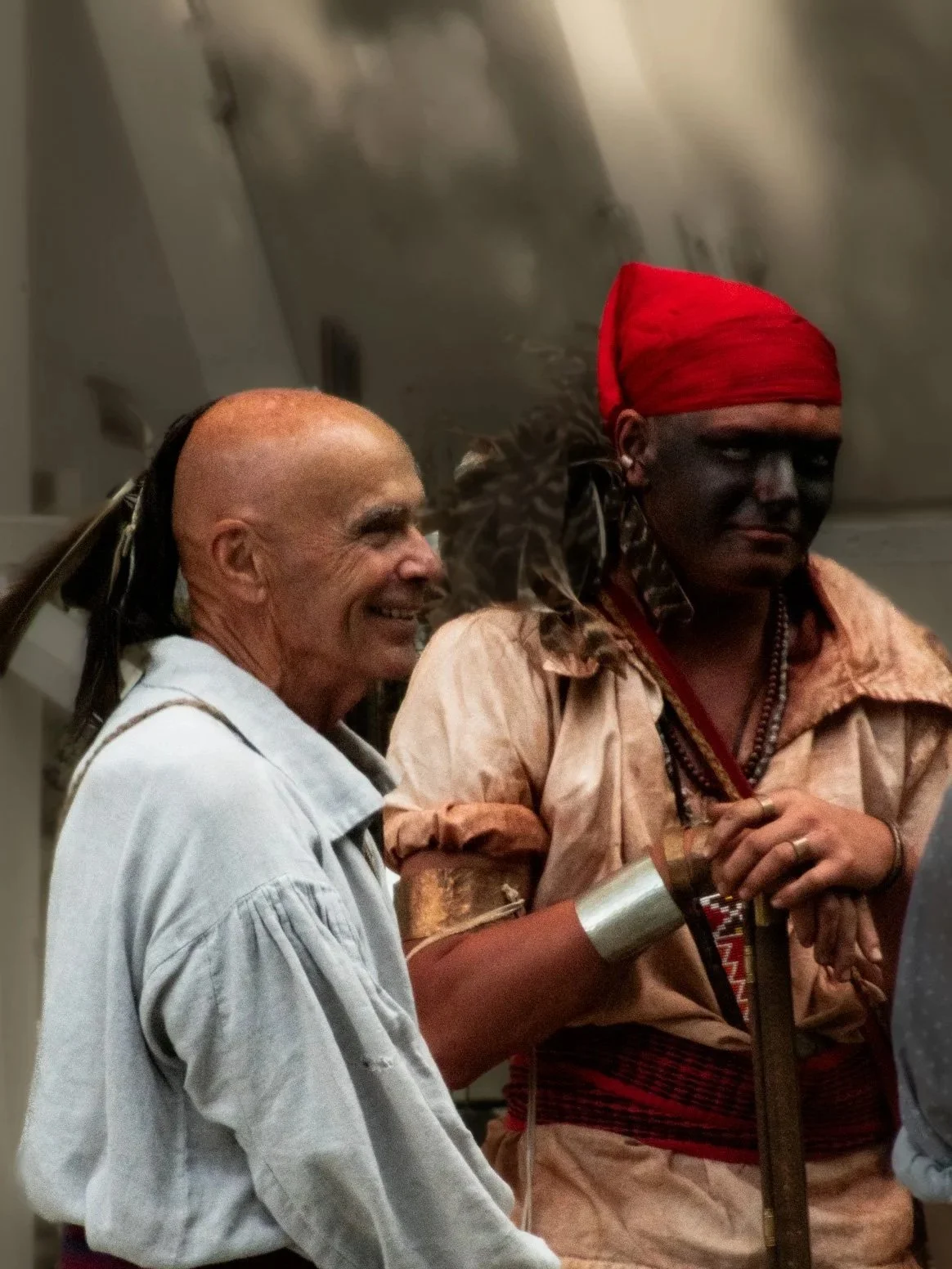 Two men, one with dark skin wearing a red headscarf and traditional attire and another with light skin with a shaved head and wearing a light-colored shirt, are standing together indoors. The man with dark skin appears serious while the other man is 