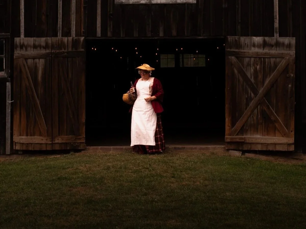 A person dressed in old-fashioned farm attire standing in front of a rustic barn doorway.