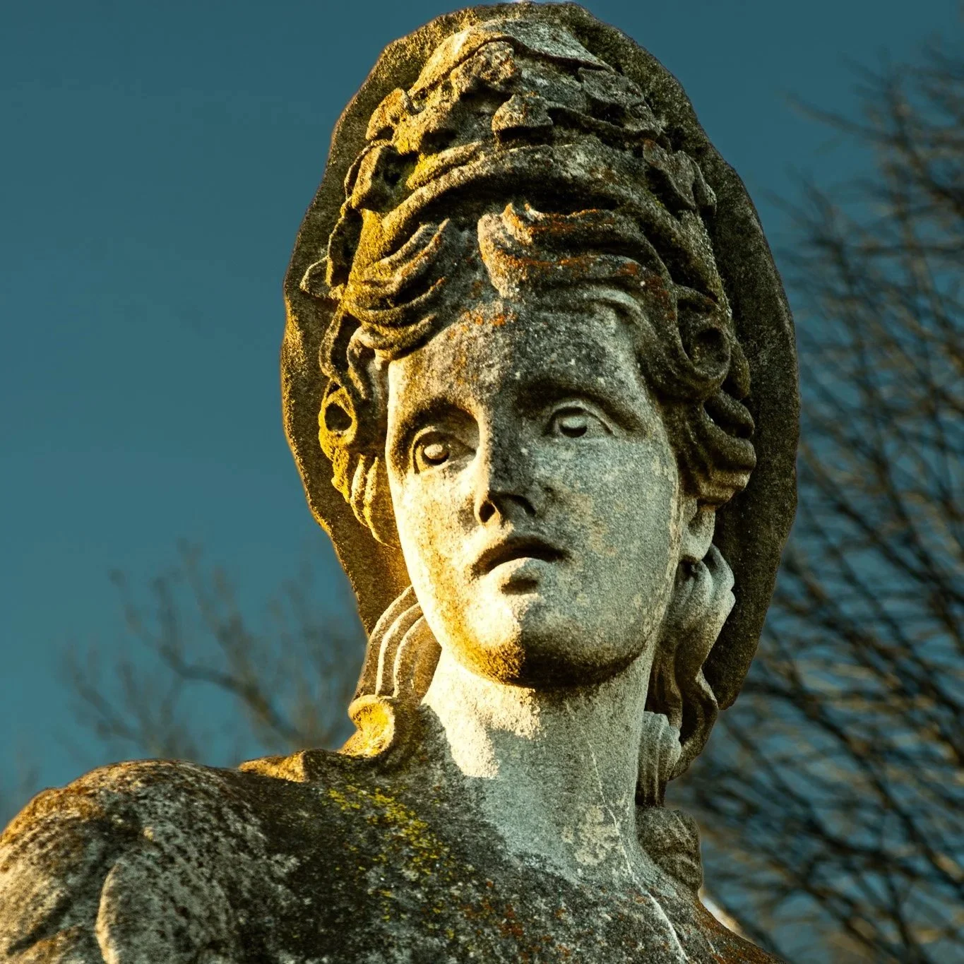 Close-up of an old weathered stone statue of a woman with an ornate headdress, with a dark sky and bare tree branches in the background.