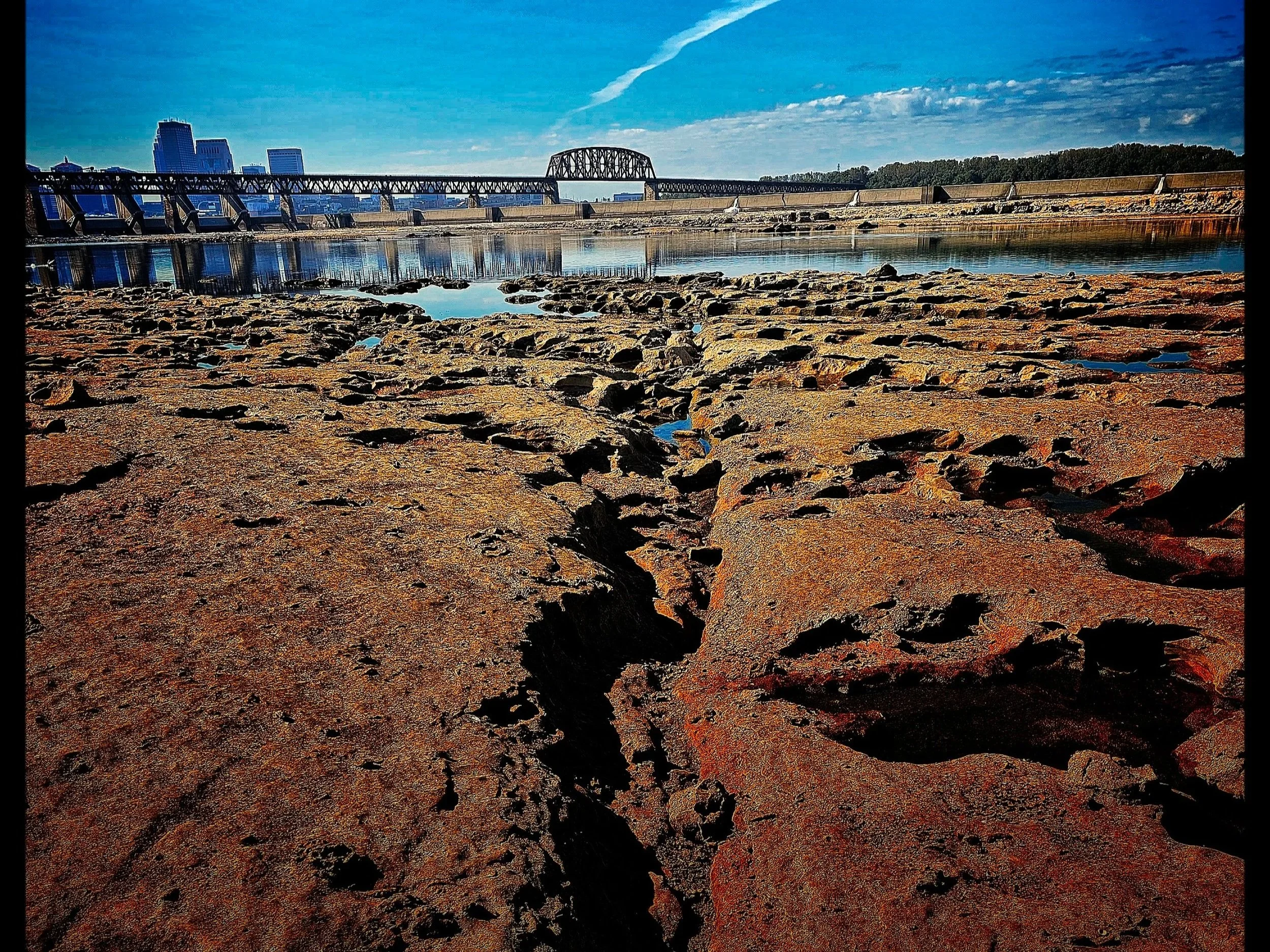 A rocky shoreline with a river, a bridge, and city buildings in the background under a blue sky.