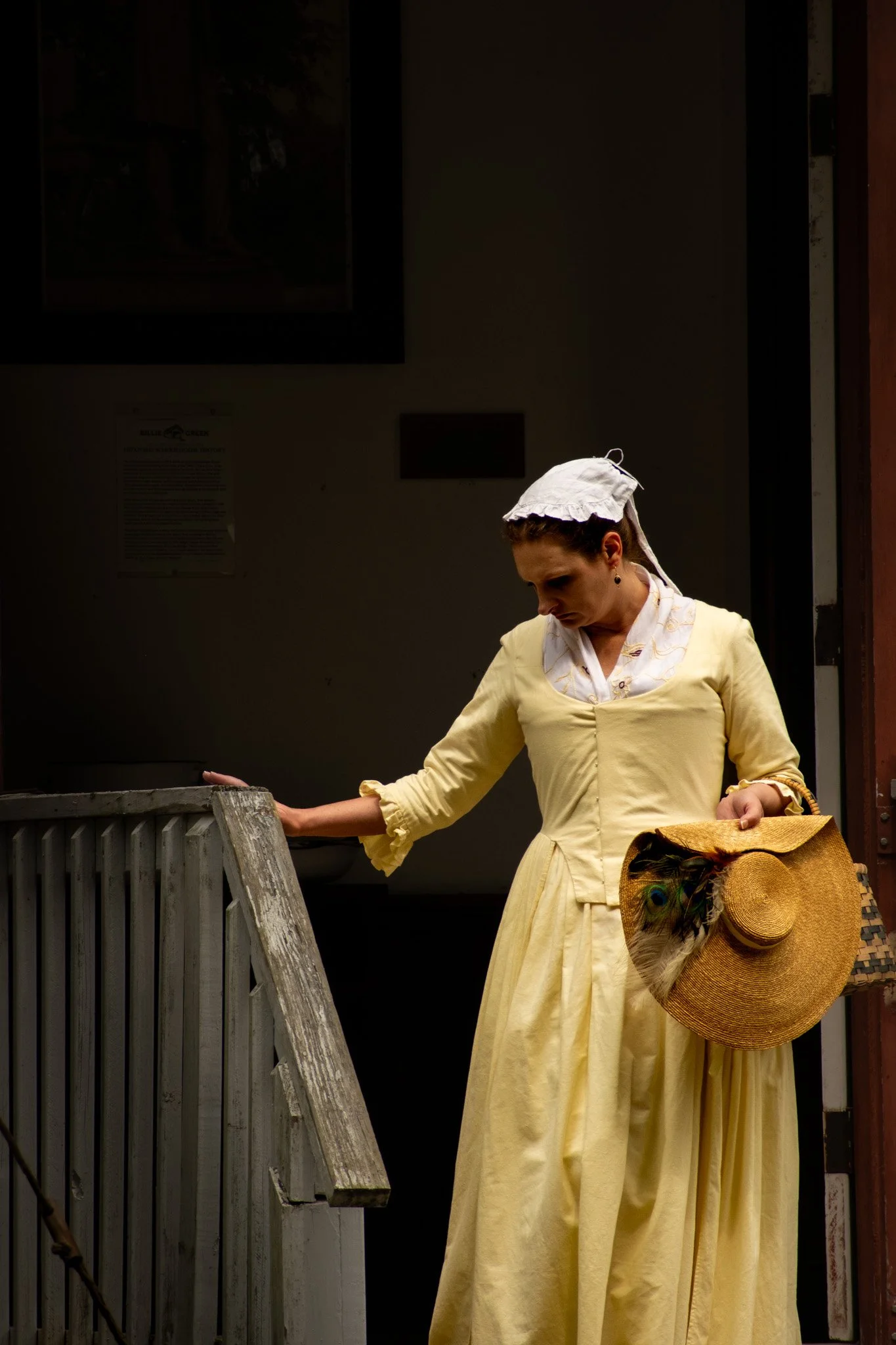 A woman wearing a yellow dress and a white head covering, holding a straw hat with a peacock feather, standing near a wooden railing.