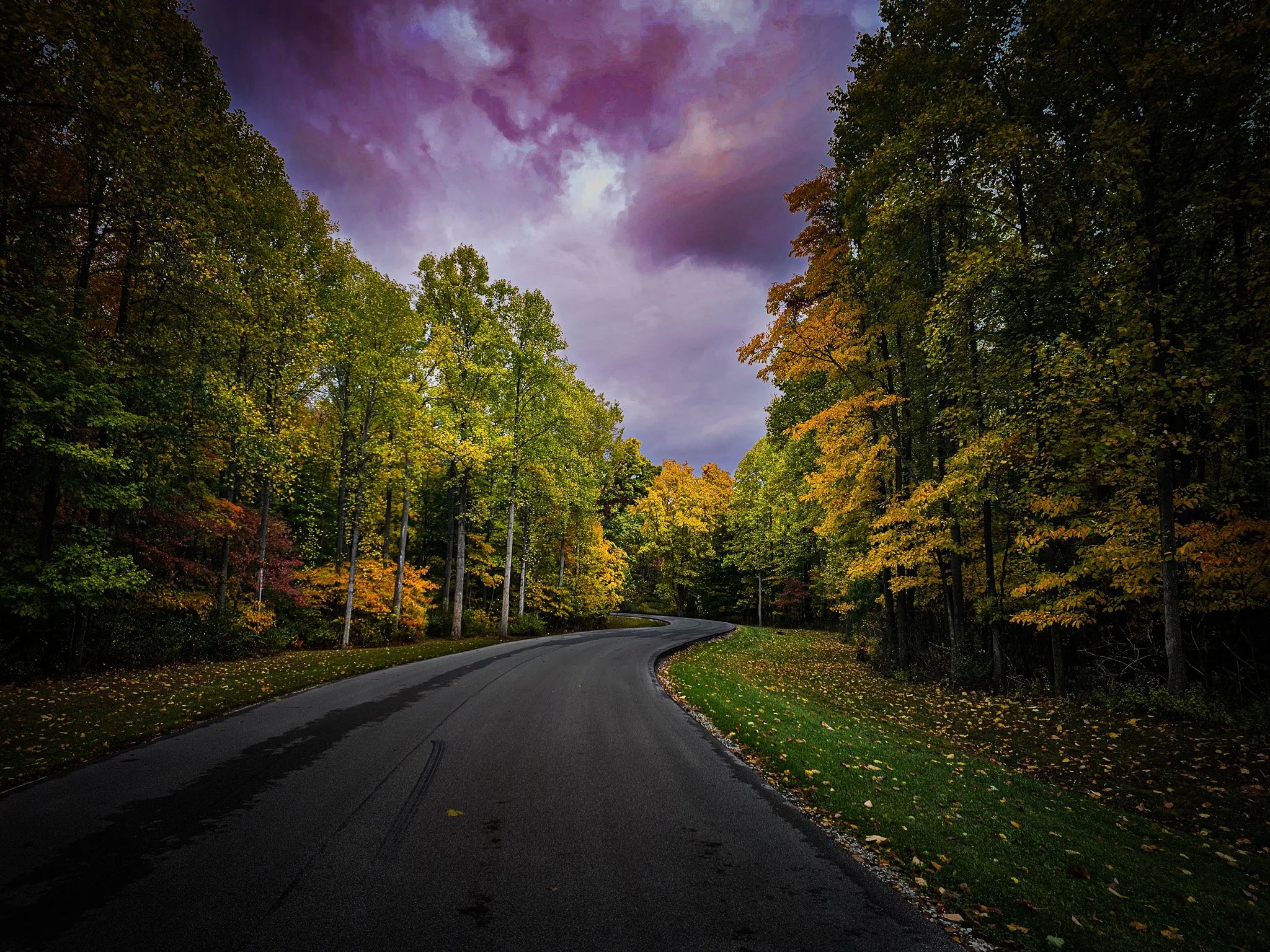 Curving road through a forest with colorful autumn foliage and a cloudy sky.