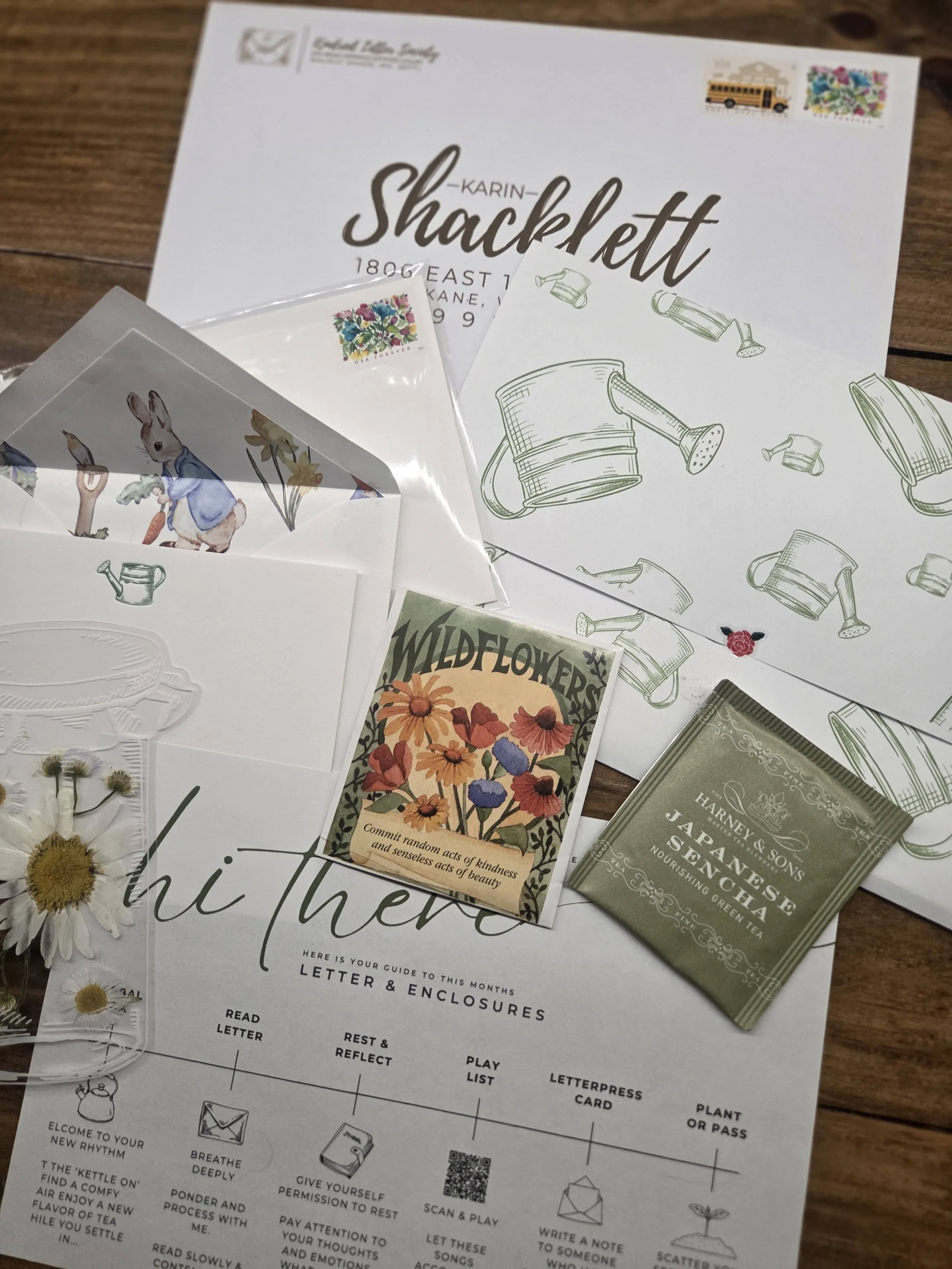 A letter from Karin Shacklett with illustrations of watering cans and garden tools, a colorful envelope, and tea packets on a wooden surface.