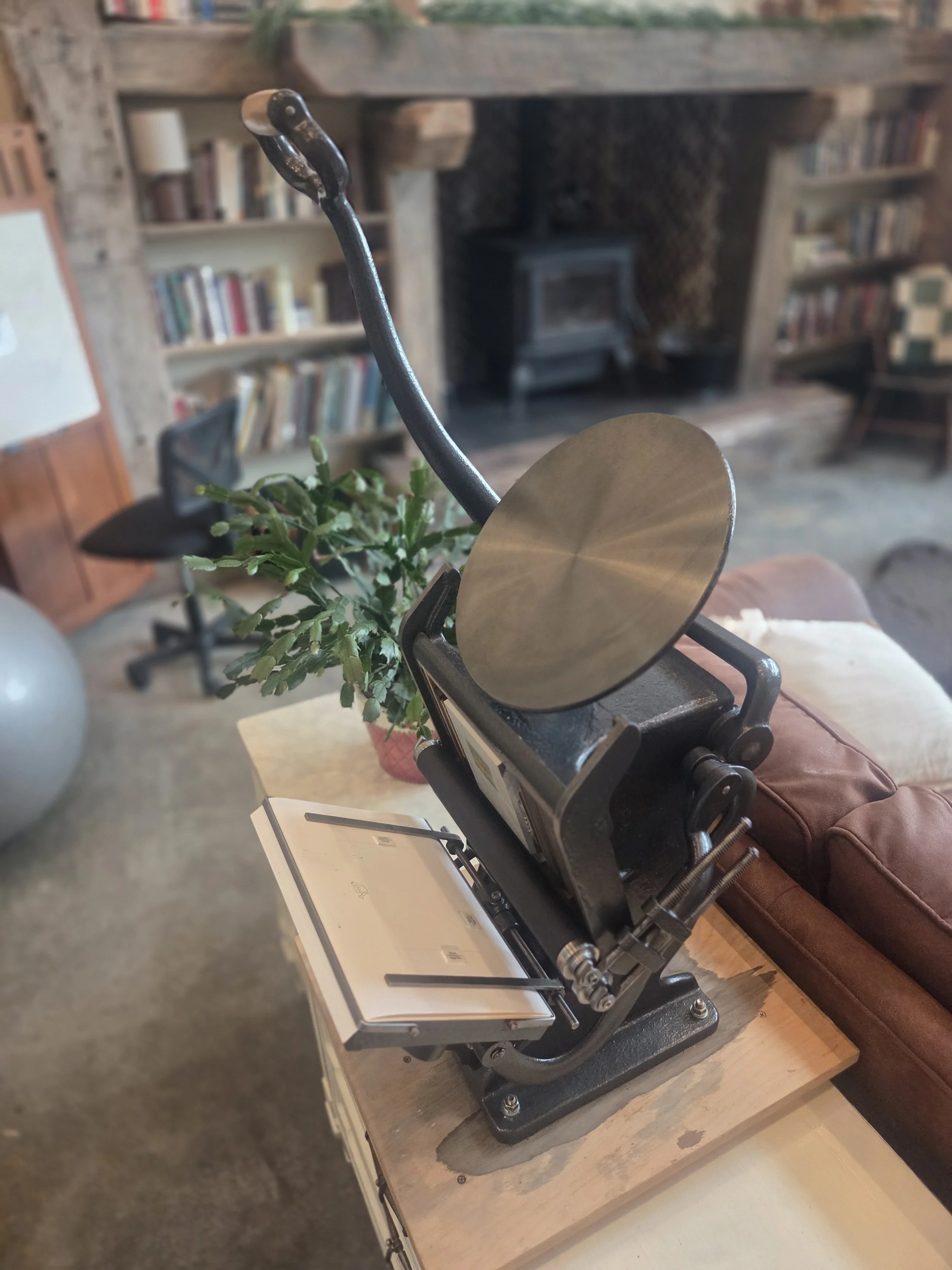 A printing press situated on a wooden table in a cozy indoor space, with a bookshelf and a small stove in the background and a potted plant nearby.