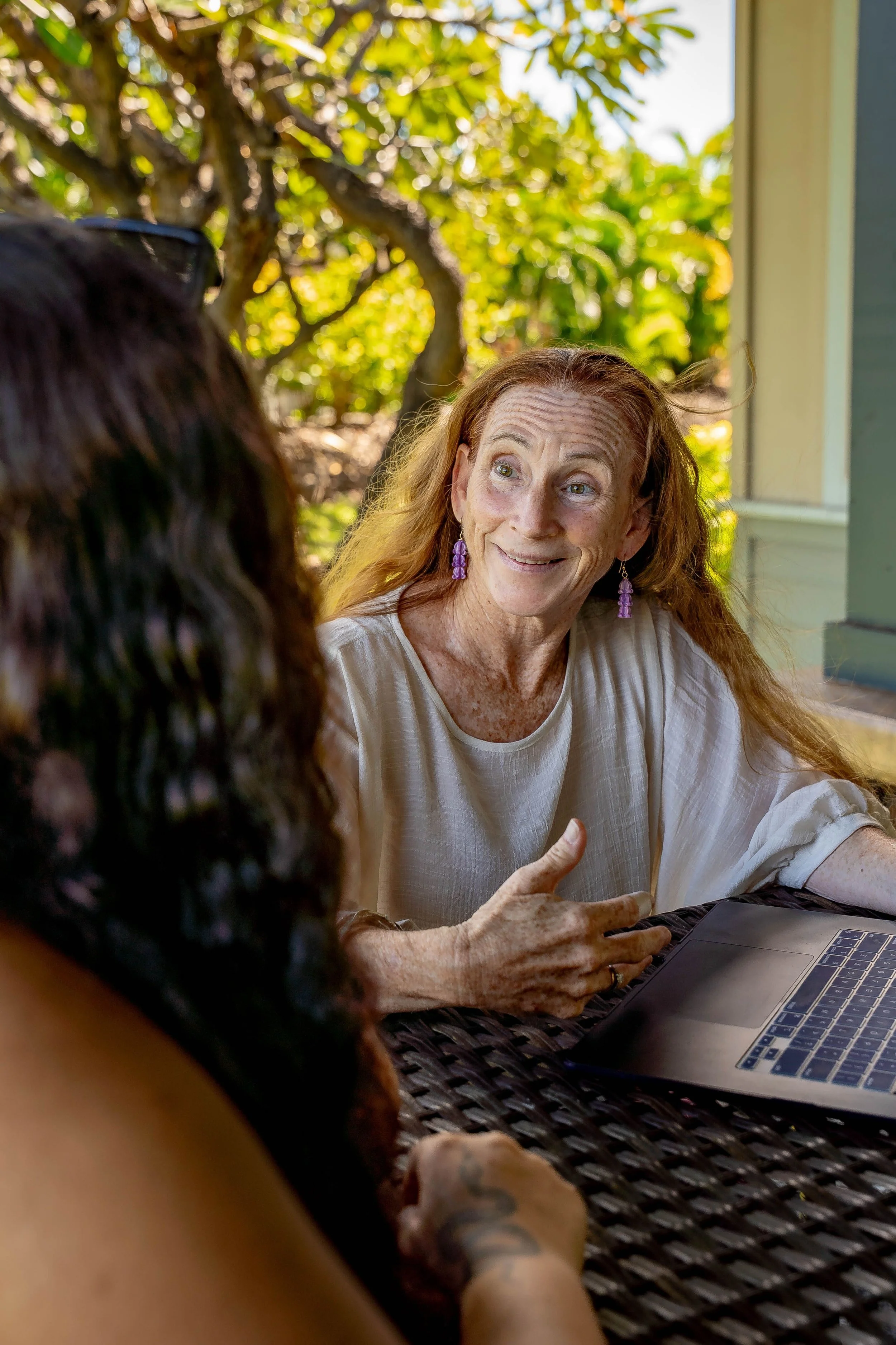 A  woman with long red hair, purple earrings, and light-colored blouse sitting at an outdoor table, talking business with another person with dark hair. There is a laptop on the table and a background of trees and a house.
