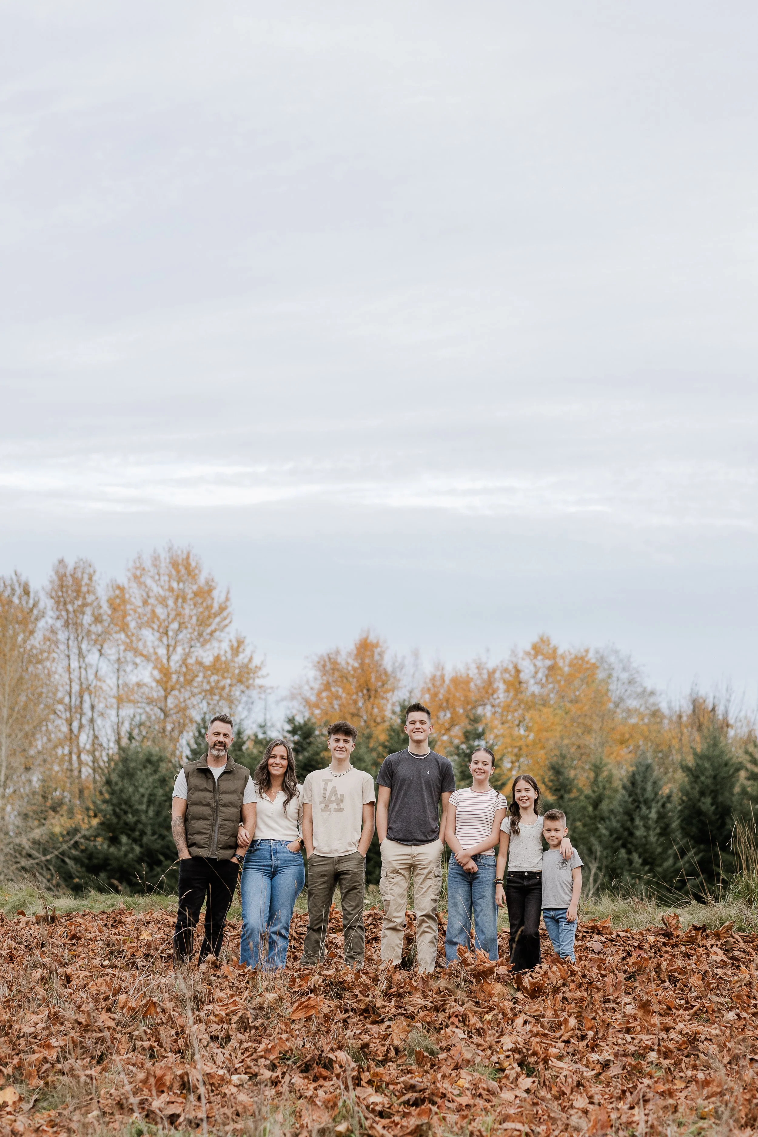 Group of eight people standing outdoors in a park during autumn, with trees with orange and green leaves in the background and a cloudy sky overhead.