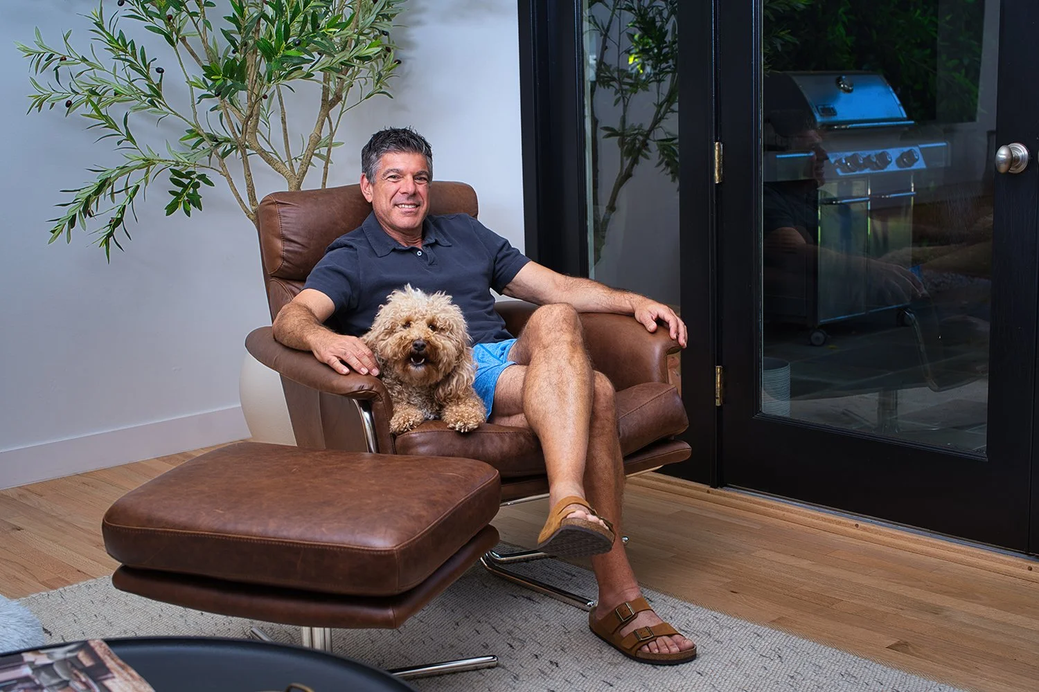 A man sitting in a brown leather chair with a small curly-haired dog on his lap, indoors near a glass door, with a large plant behind him, and a wooden floor.