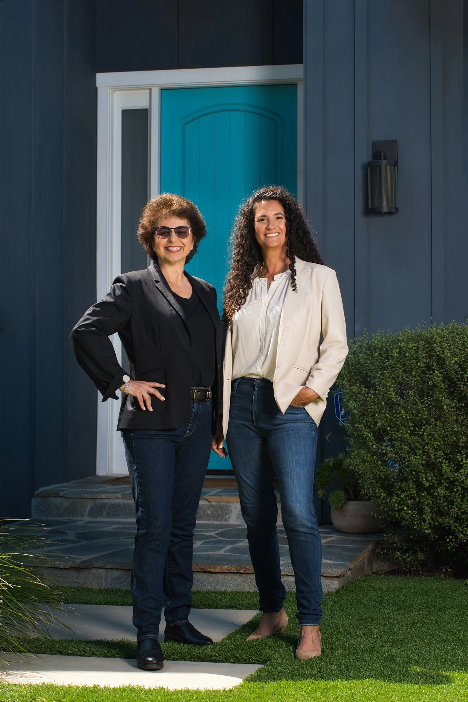 Two women standing outside a house, smiling. One woman has curly hair and is wearing a white blazer; the other woman has short curly hair and is wearing sunglasses and a black blazer.