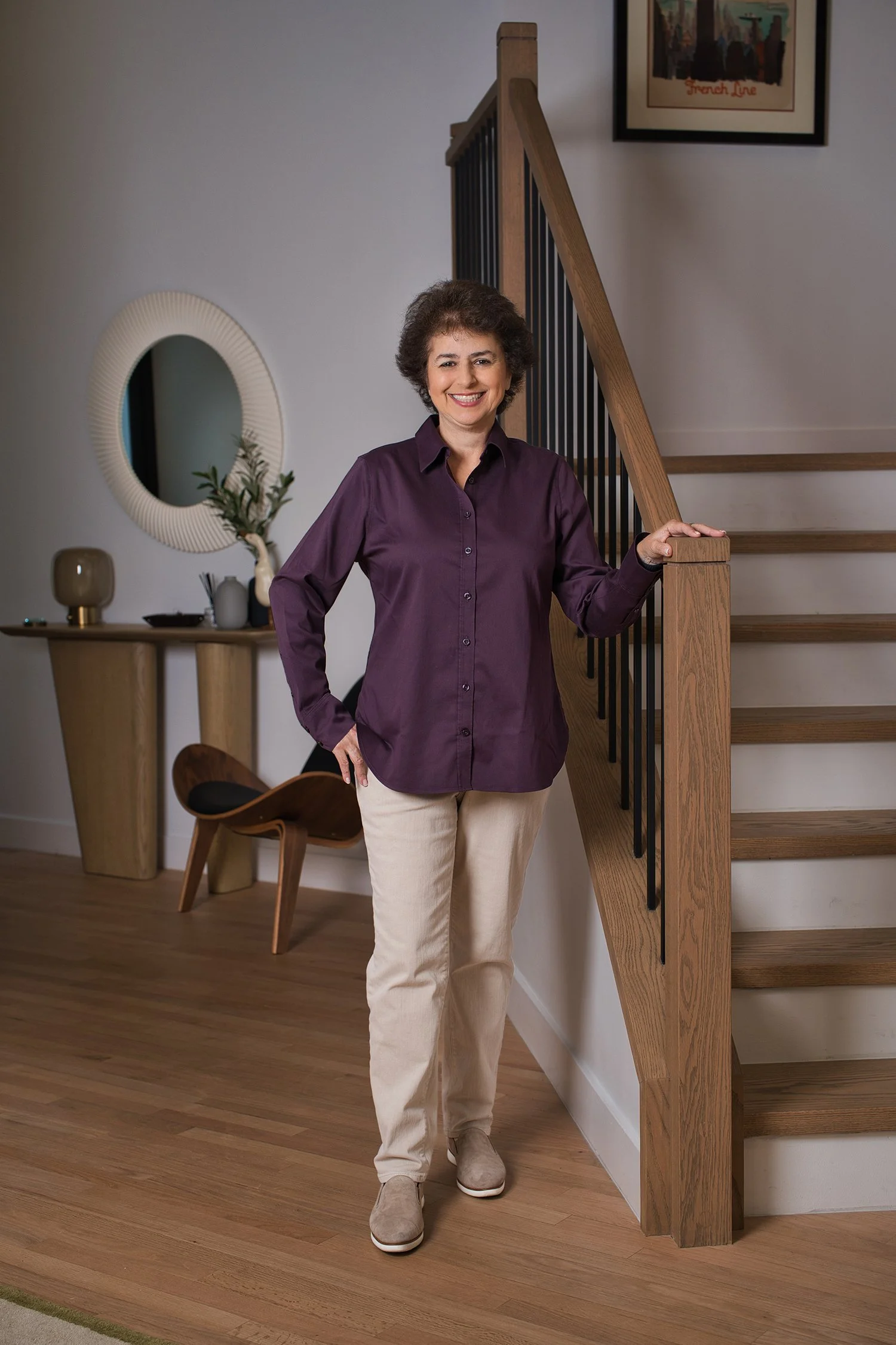 Woman standing on hardwood floor by staircase with wooden banister, smiling and wearing a purple shirt, beige pants, and beige shoes. There is a side table with decorative items, a mirror, and a framed picture on the wall in a well-lit interior.