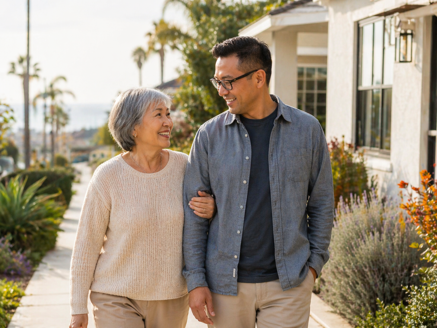 An elderly woman and a middle-aged man walking arm in arm outside a house, smiling at each other on a sunny day with palm trees in the background.