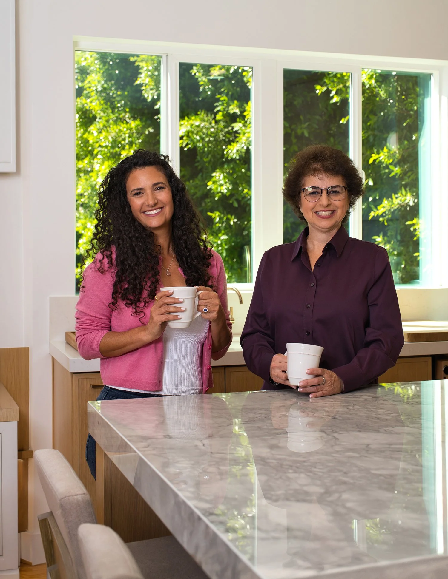 Two women standing in a bright kitchen behind a marble countertop, holding white mugs, with a large window showing green foliage outside.