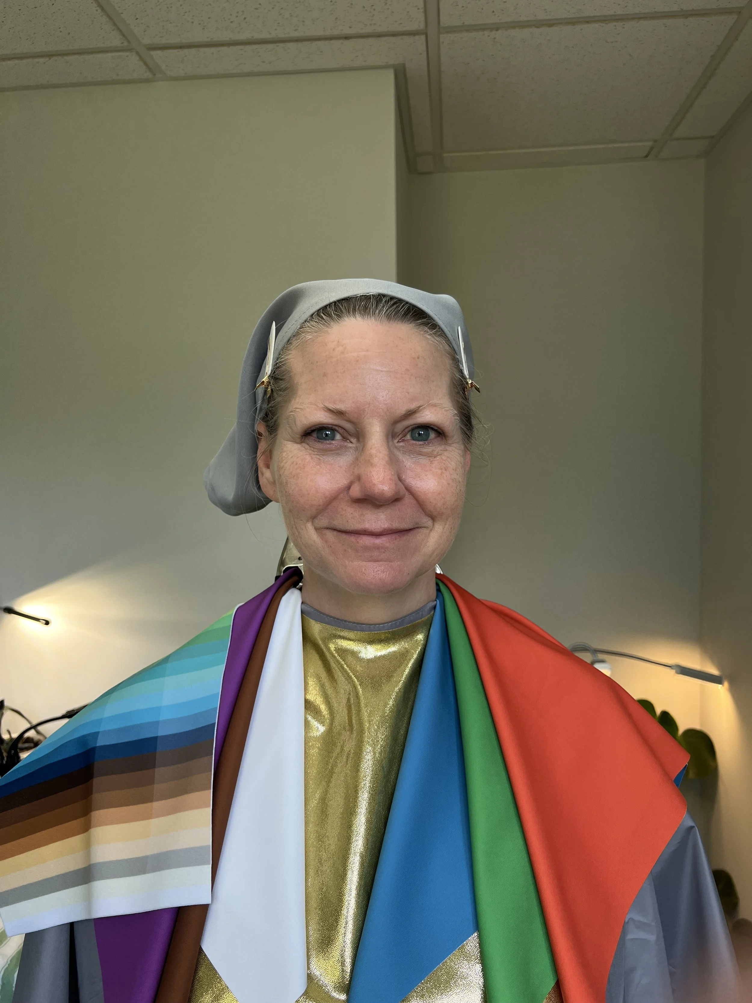 A woman with light hair, wearing a silver head covering, smiling, and draped in a rainbow flag with multiple colors including purple, white, gold, blue, green, and red.